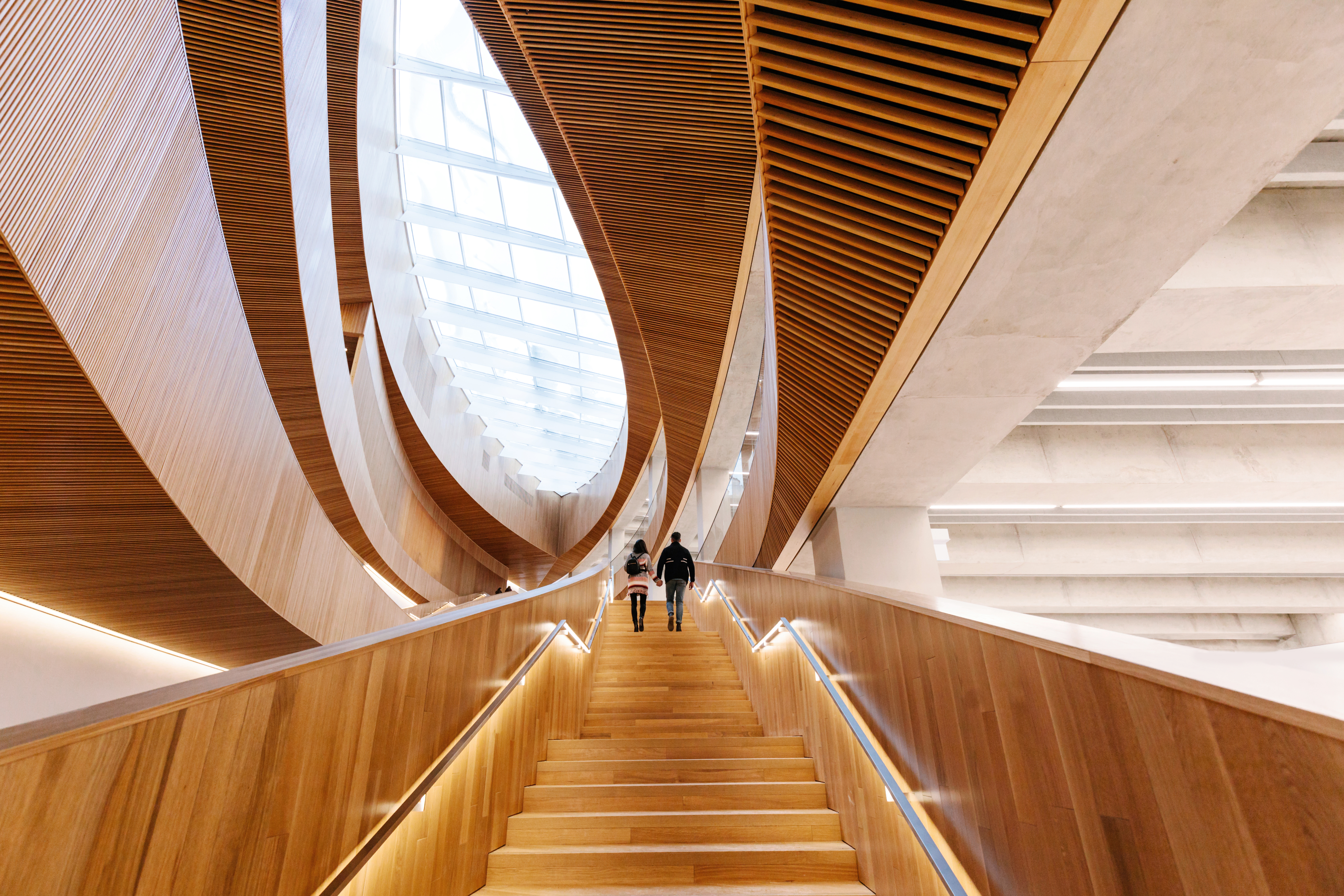 Couple walk up wooden entrance staircase of Central Library with modern geometrical façade leading to upper floors and atrium