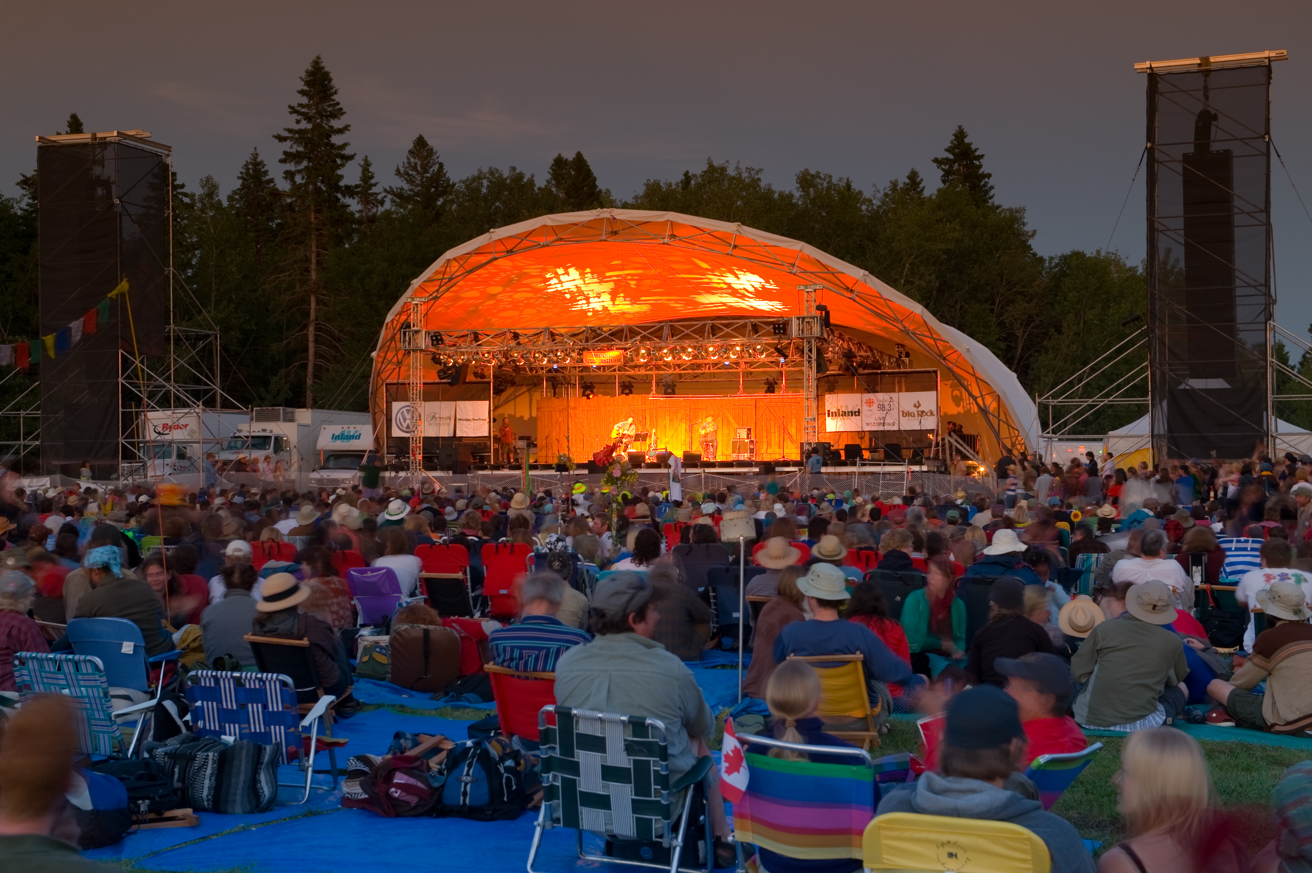 People sit on fold up chairs and enjoy picnics in front of an outdoor music stage