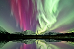 Pink, purple and green colours of the Northern Lights above mountains and reflected in a lake