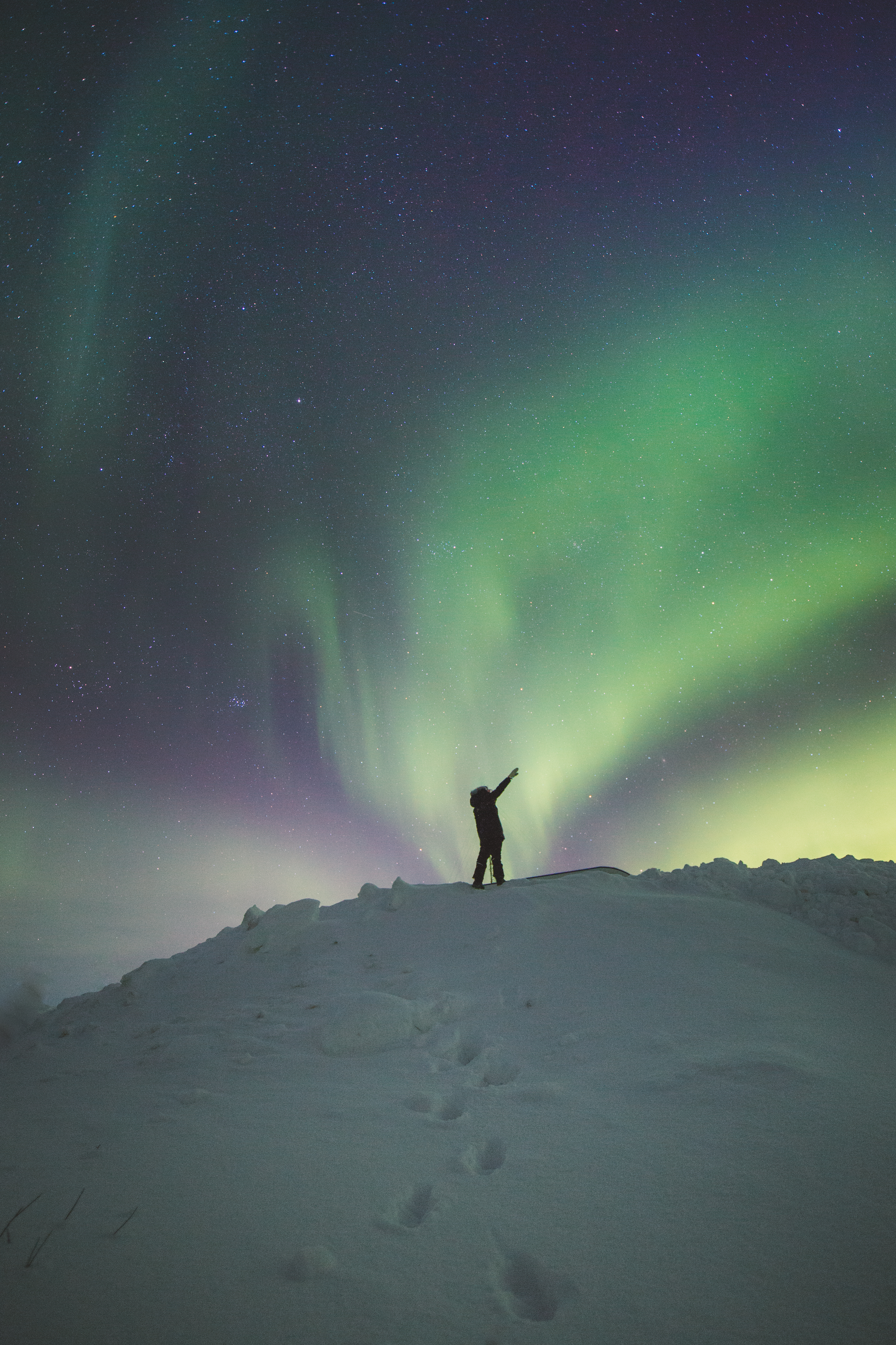 A person standing on snowy ground beneath pristine colourful Northern Lights with their arms reaching upwards and in the air 