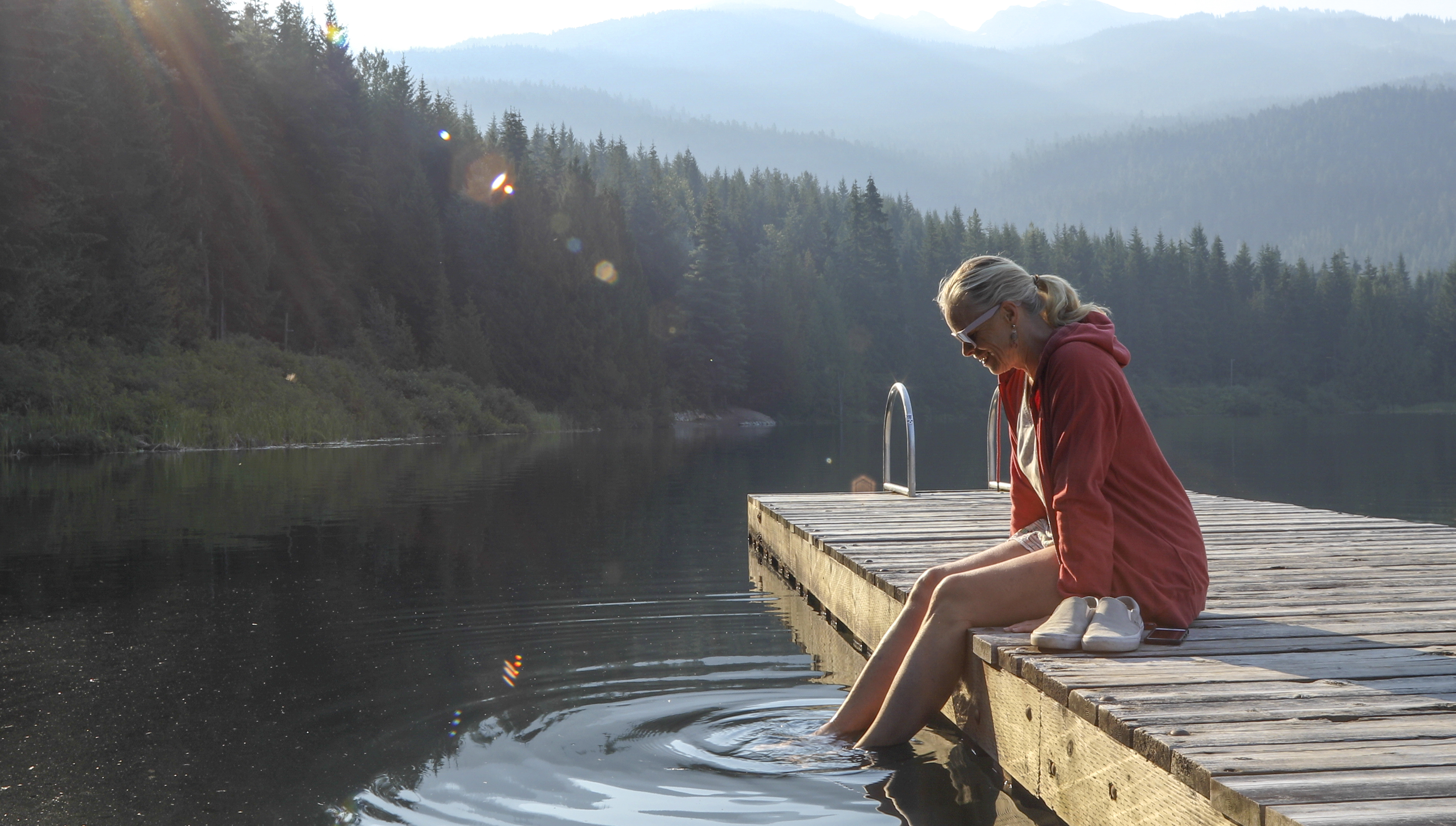 Senior woman sitting on wooden dock soaks feet in Lost Lake, Whistler, near Blackcomb village
