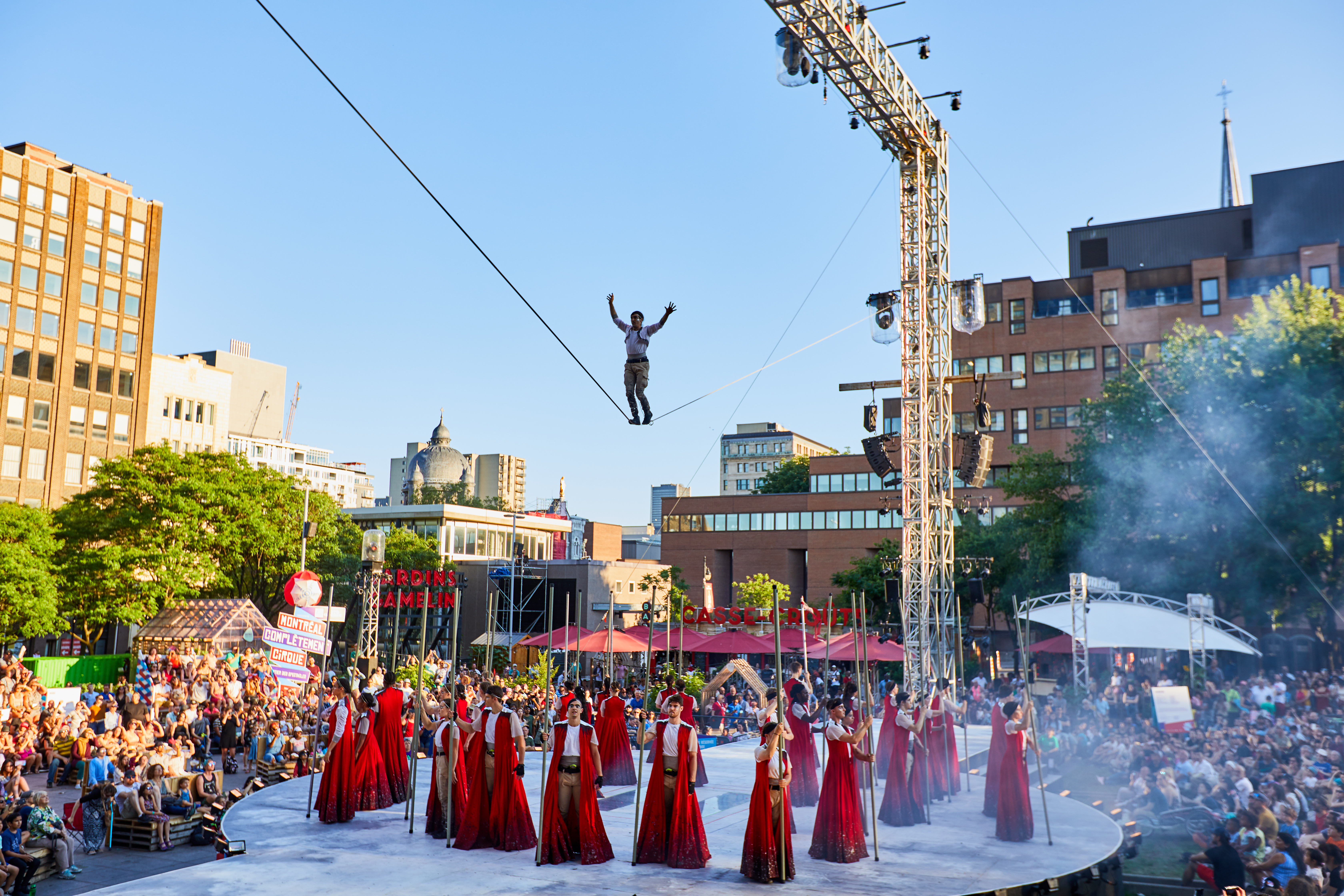 A slack line performer above a group of circus artists on an outdoor stage in front of a crowd