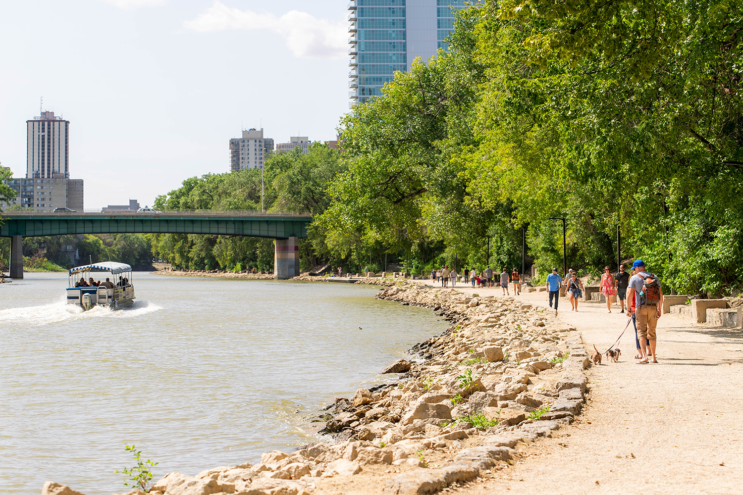 People walk along a curving river trail at The Forks as a boat cruises on the water