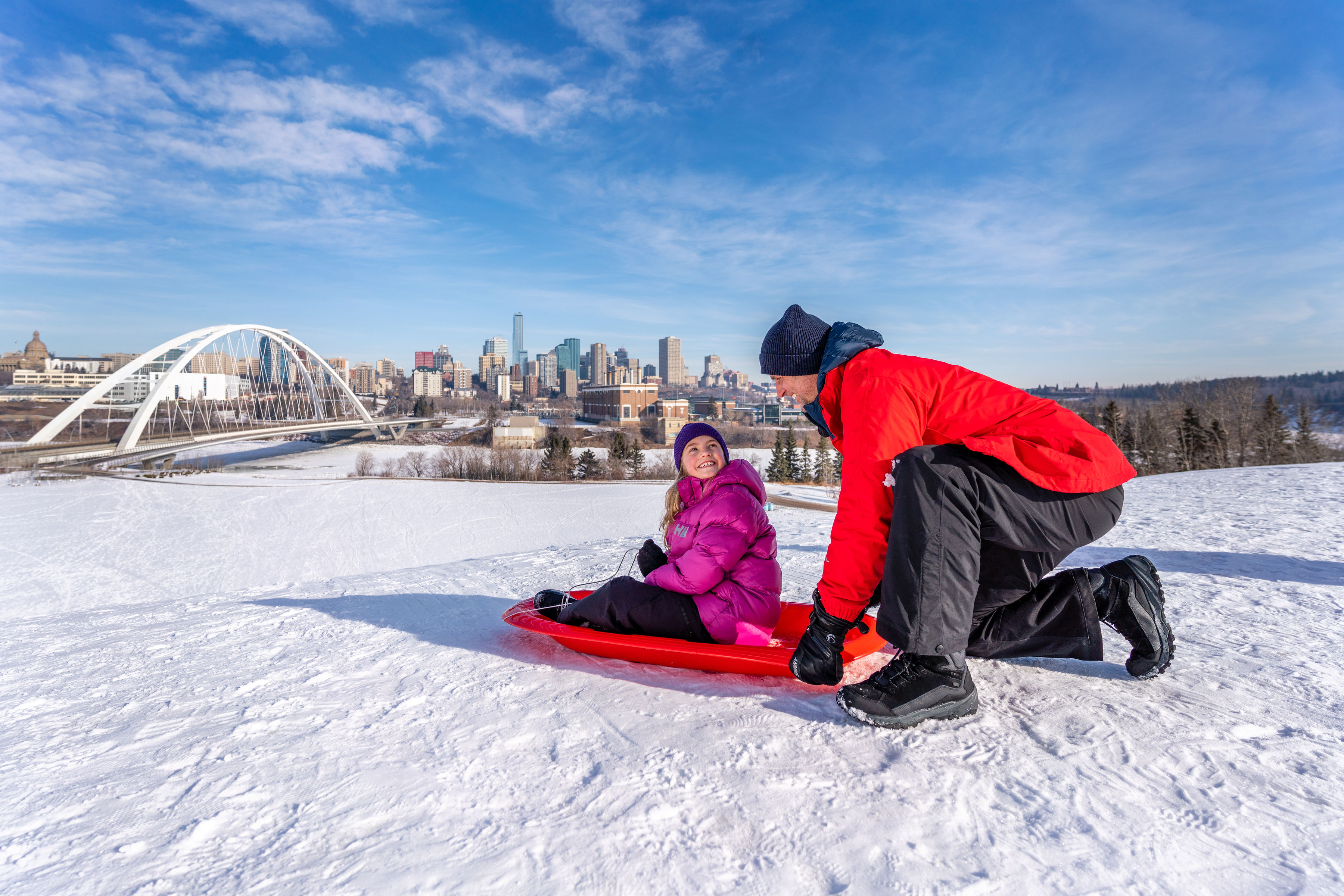 A dad helping a child on a toboggan with view of Edmonton city in the distance