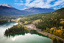 The Rocky Mountaineer train travels along a lakeside, fringed with fall colors with a mountain range behind.