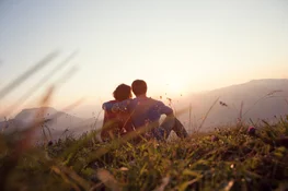 A couple sitting on a grassy slope stares out at the sunset