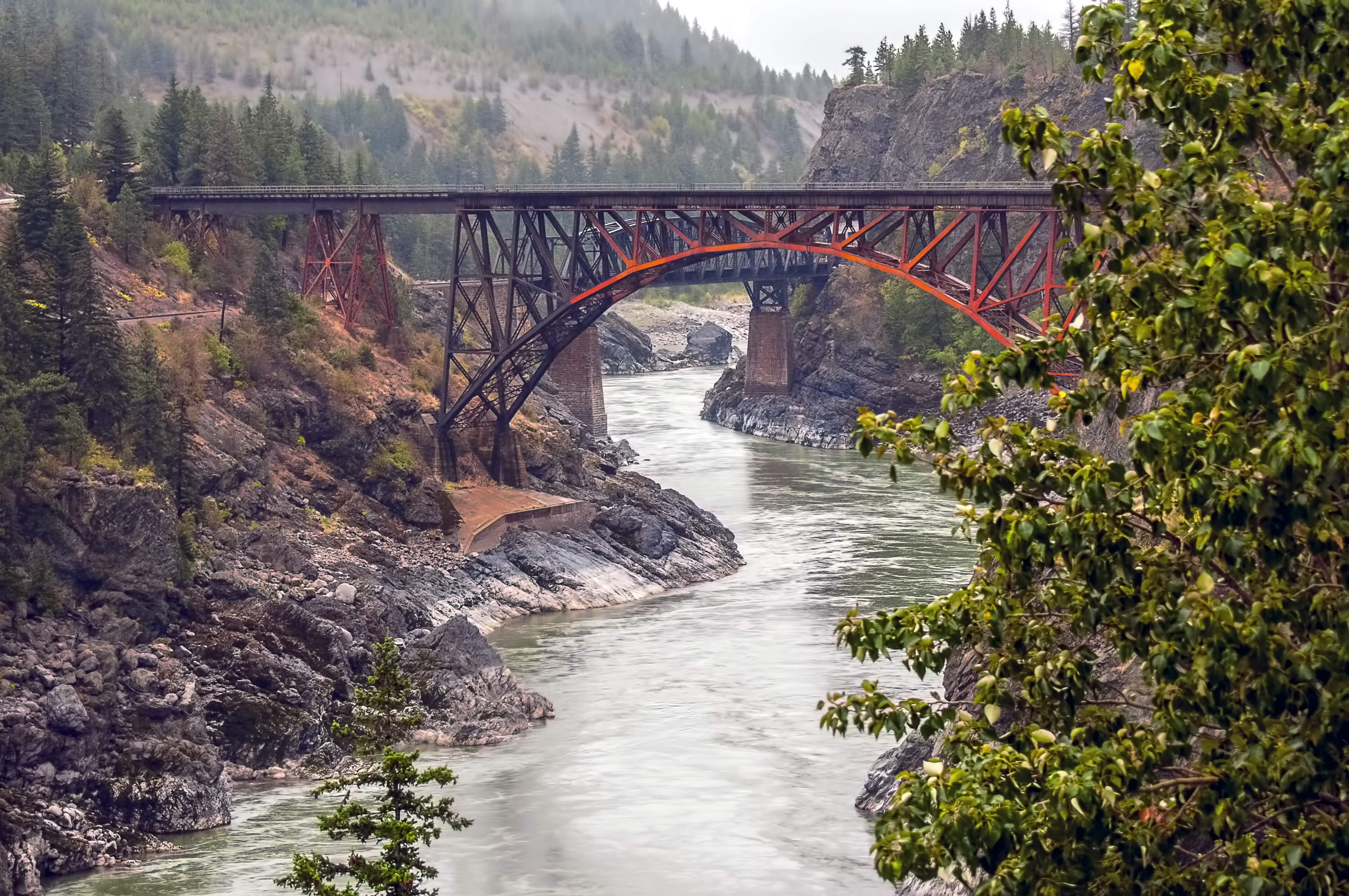 A rail bridge crosses the Fraser River in a misty landscape