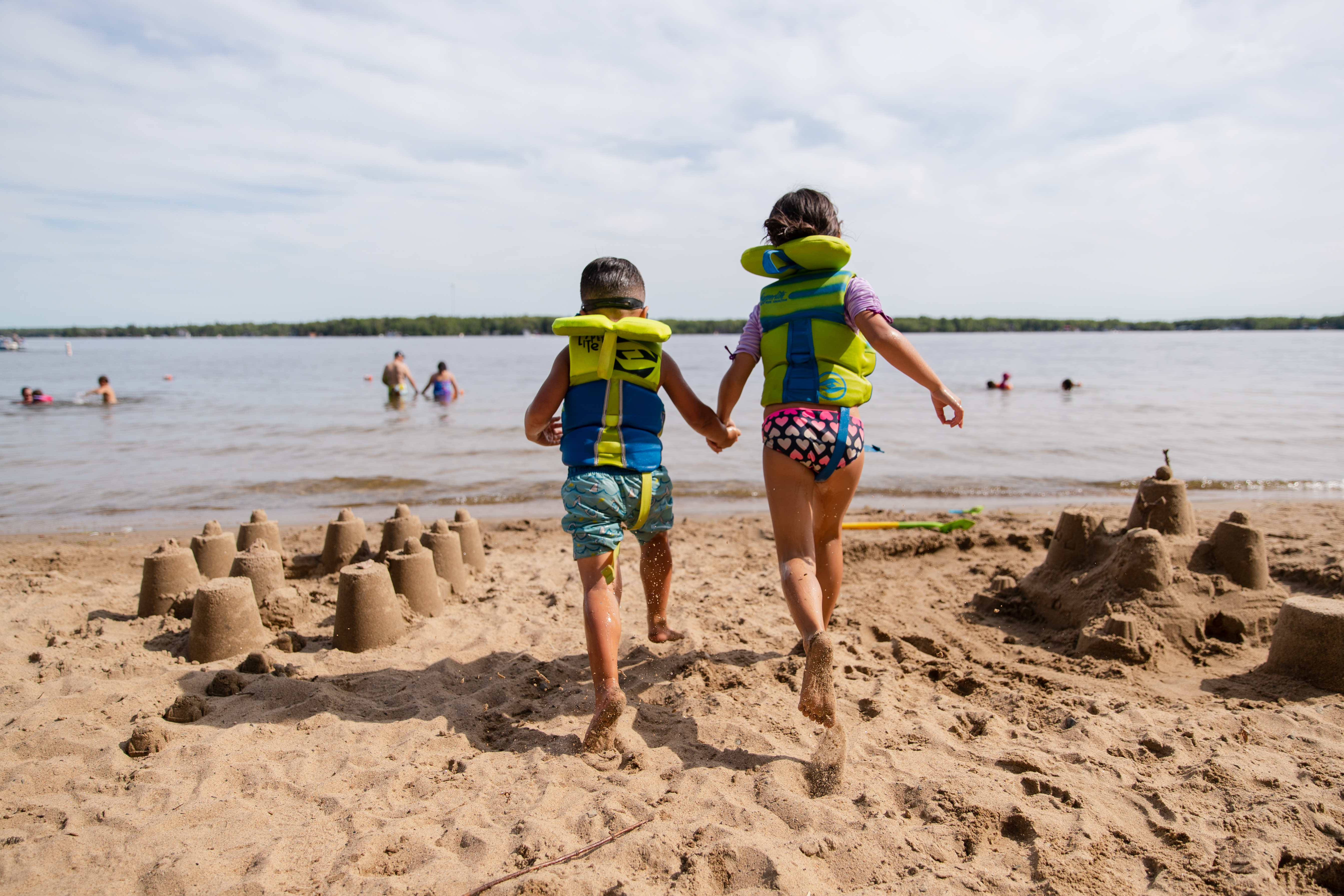 Two children wear lifejackets and hold hands as they run past sandcastles toward a lake