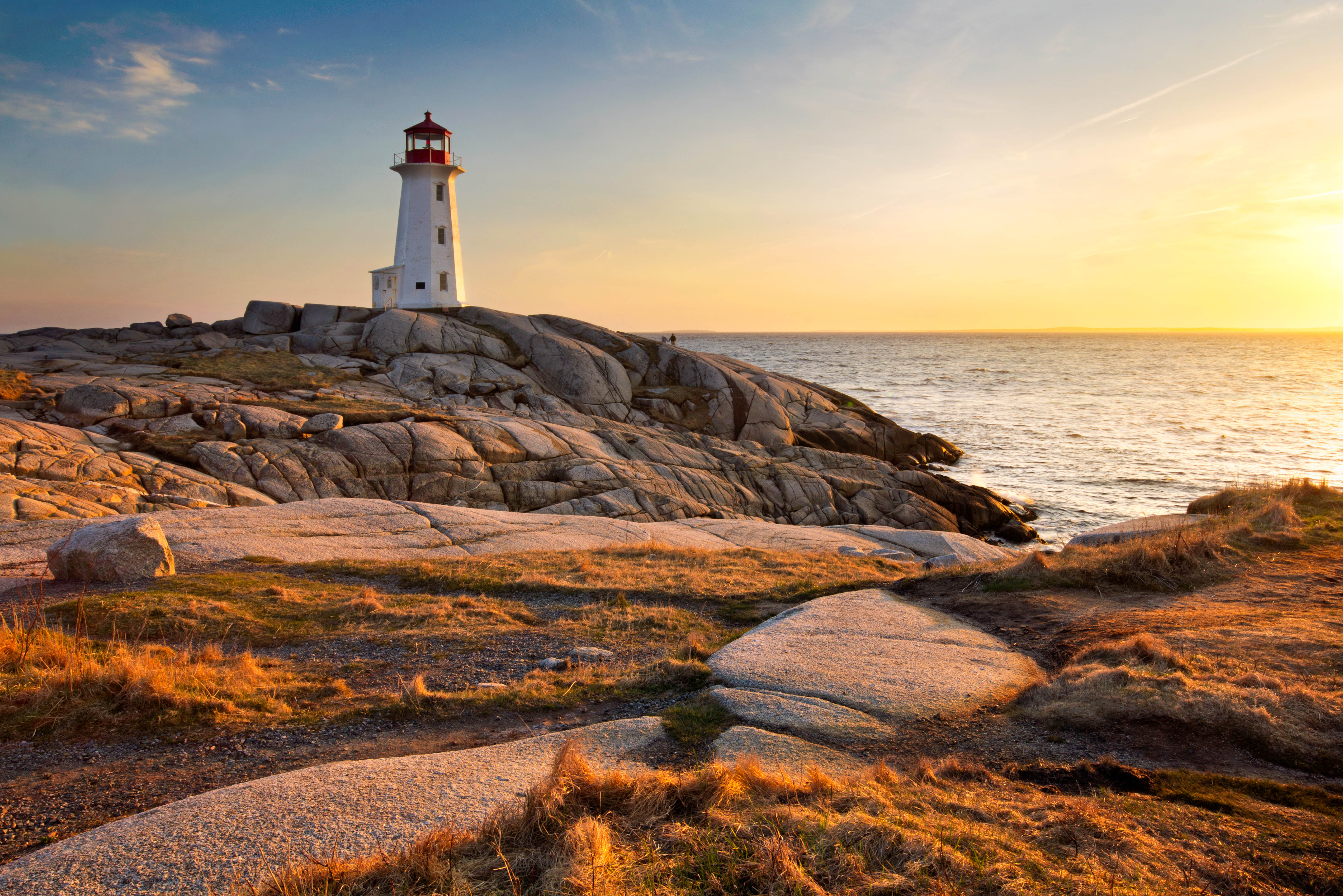 Lighthouse on rocky shoreline at sunset with an orange-blue sky in the background..