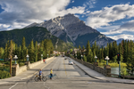 Two people with bikes cross the road in Banff with mountains in the background