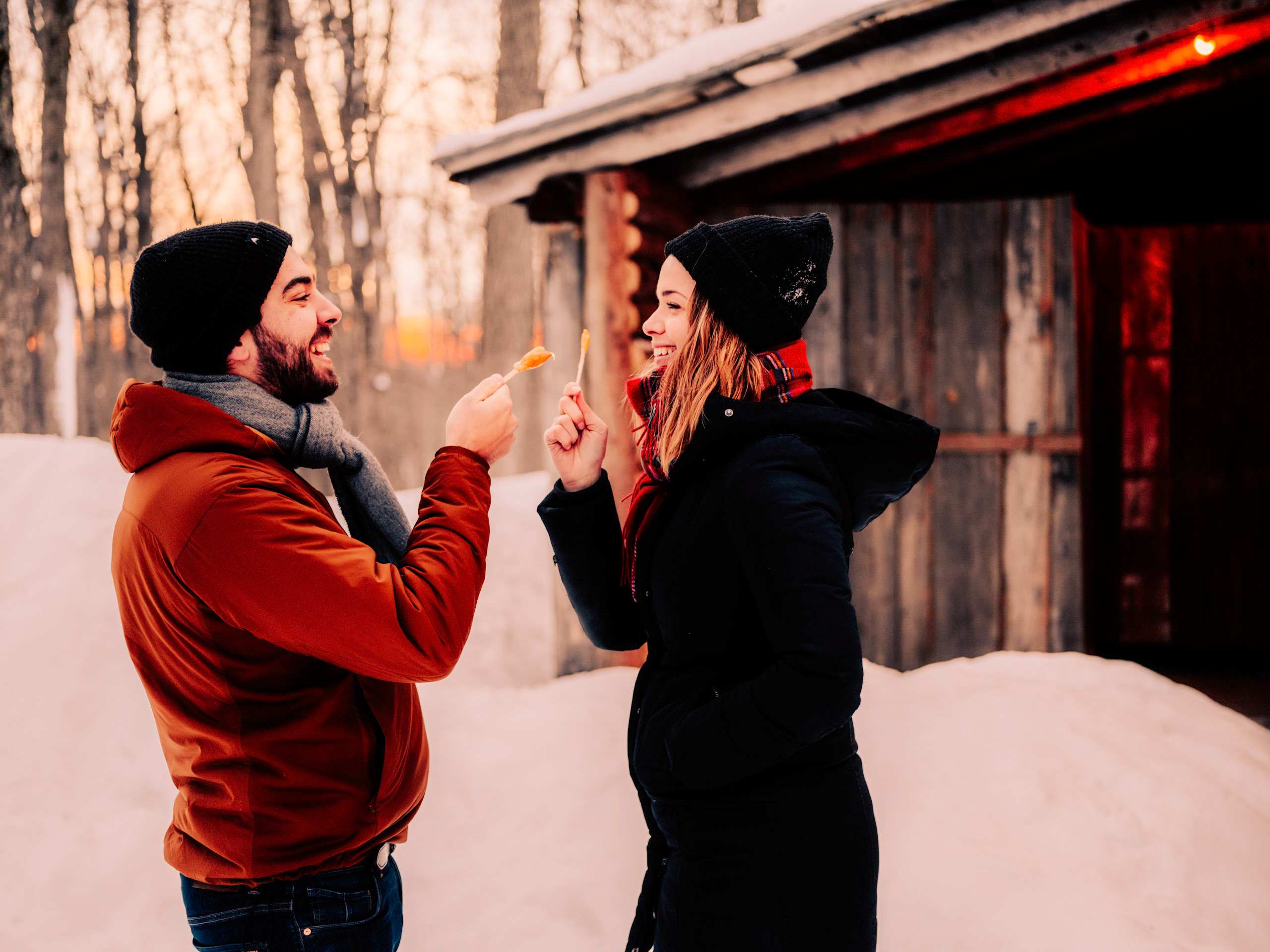 Couple smile and pose with maple sap lollipops outside of sugar shack, La Sucrerie de la Montagne, surrounded by maple trees after snow