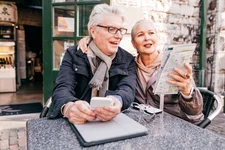 Senior couple resting in outdoor coffee shop, man holds phone, woman holds newspaper
