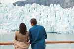A couple stand on the deck of an Alaska cruise ship looking out at an iceberg