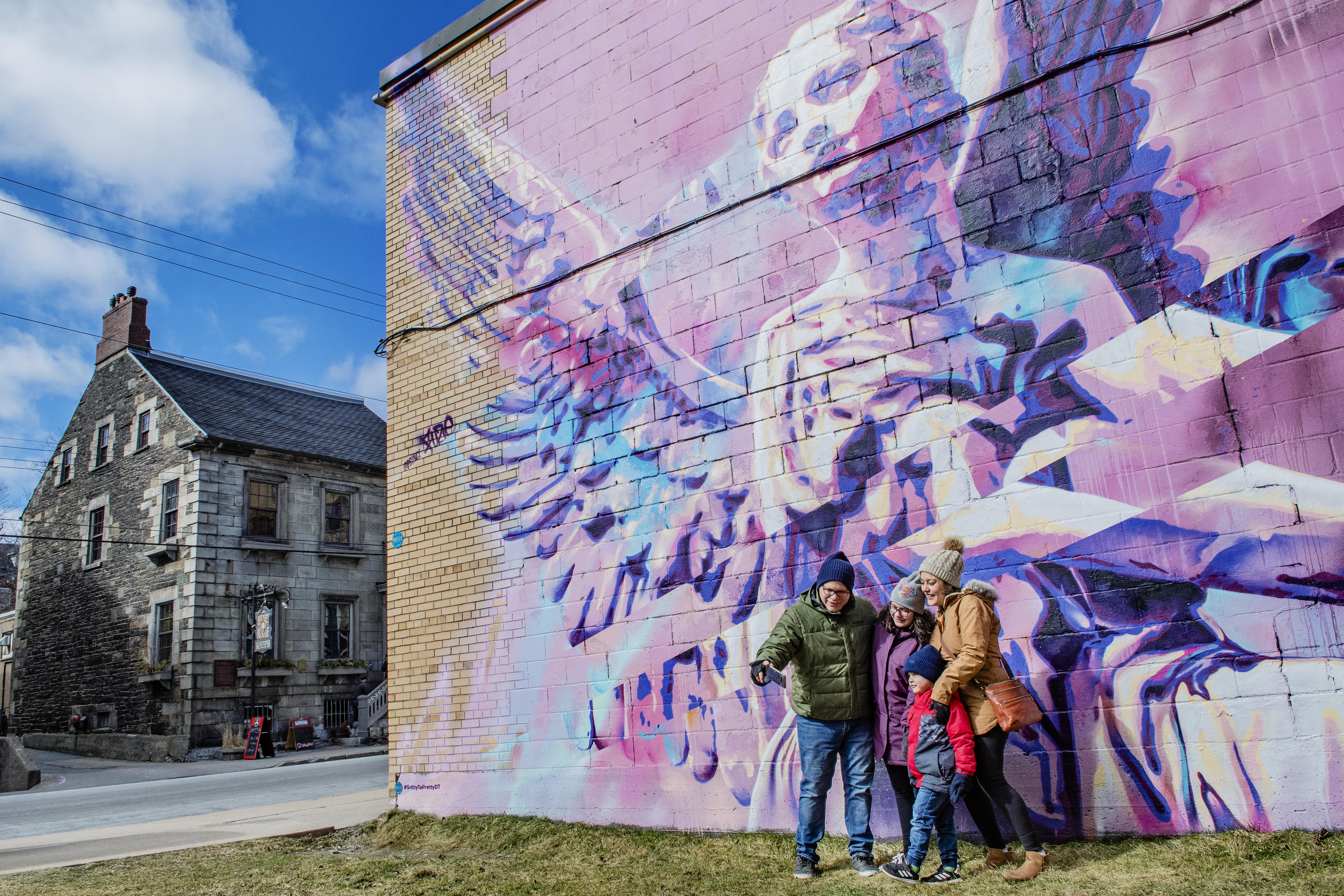 A family standing against a brick building which has a purple art mural of an angel on it