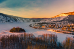 Blue and light orange sky casts its warm colours on a snowy Dawson City and its buildings, forest and mountains during winter