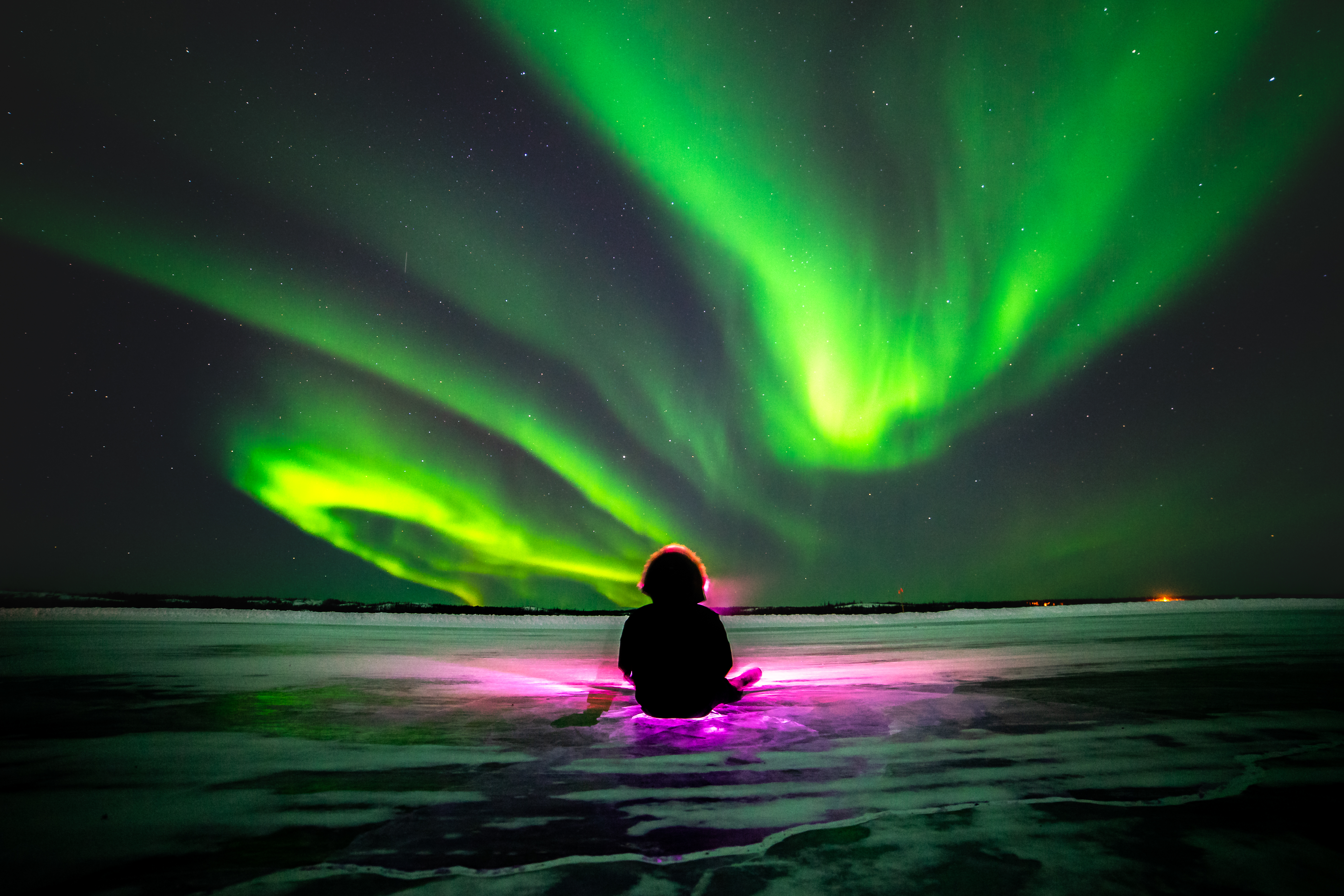 Person sits on ice while viewing Northern Lights glow revealing dark sky and its stars in the capital of the Northwest Territories in Canada