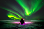 Person sits on ice while viewing Northern Lights glow revealing dark sky and its stars in the capital of the Northwest Territories in Canada