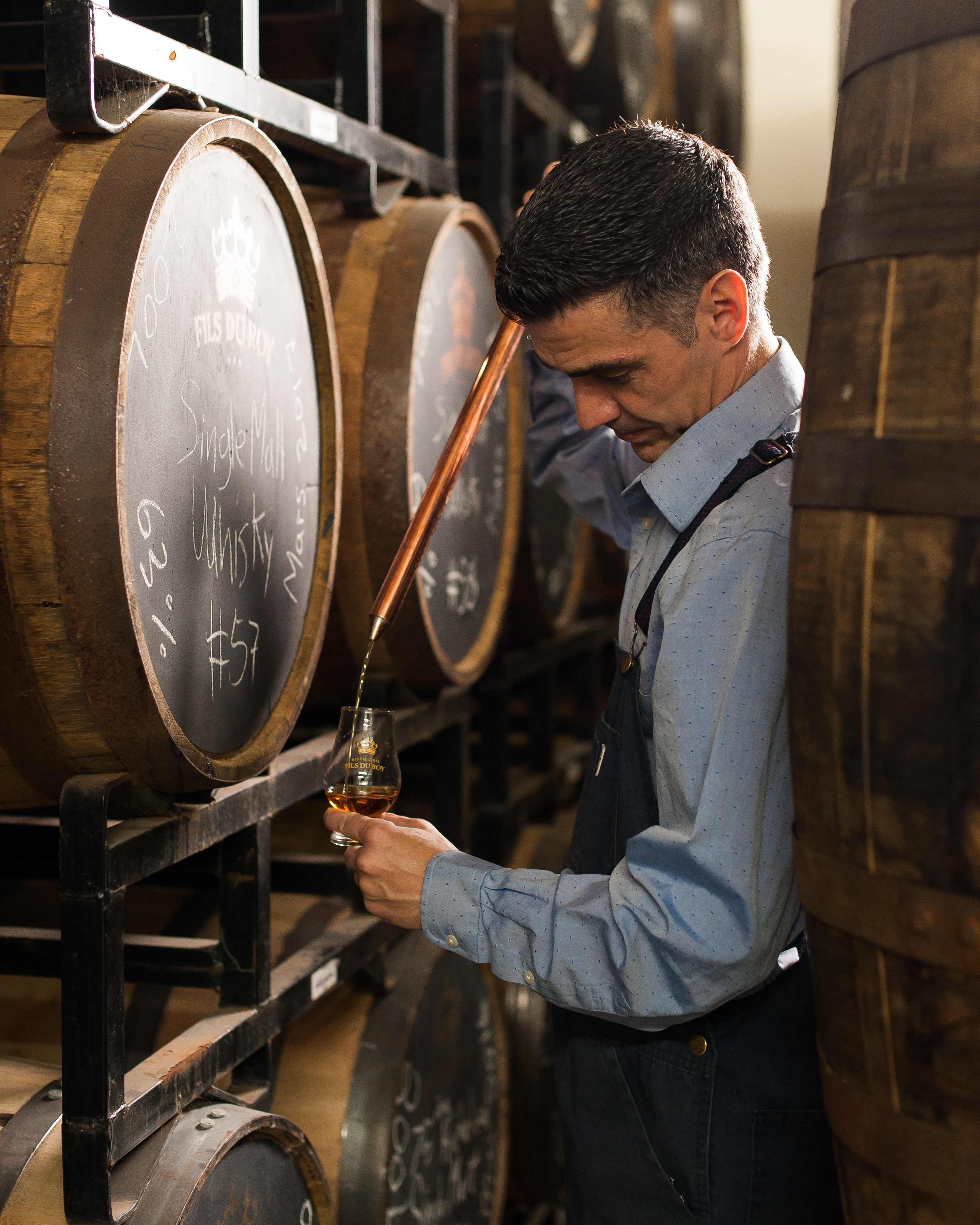 Distiller extracts small portion of spirit produced with local ingredients from aging barrel for sampling using glass thief at Artisanal Acadian distillery, Distillerie Fils du Roy