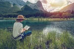 Woman reading by a lake with mountains in background