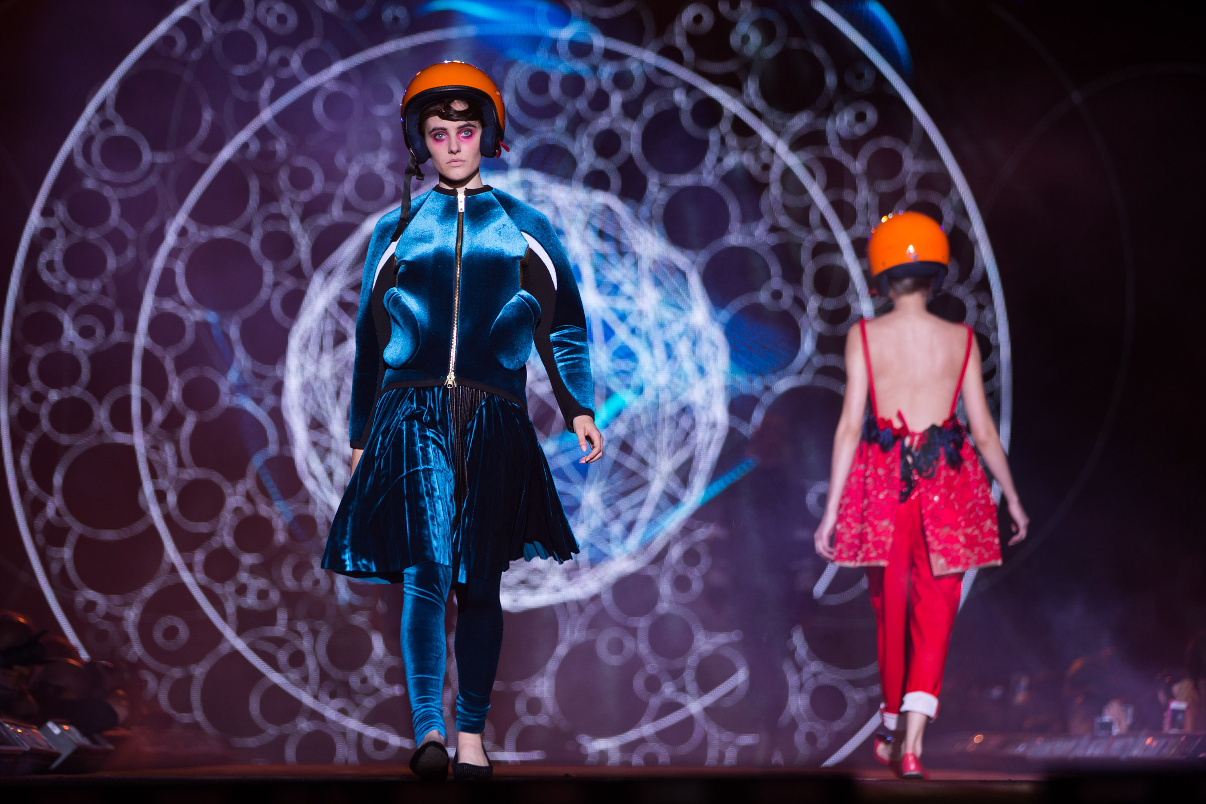 A model wearing a blue velvet outfit and orange helmet walks on a catwalk