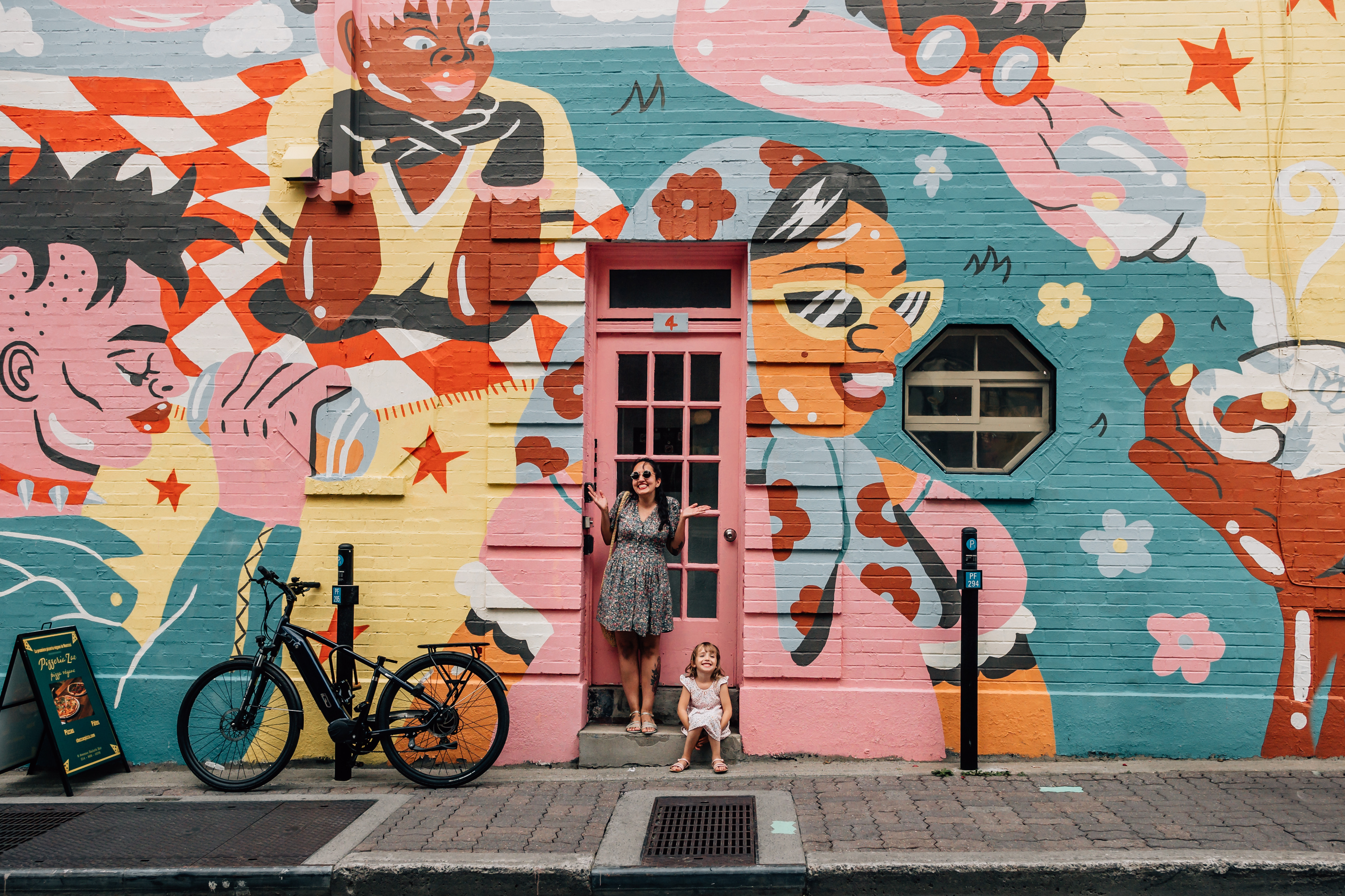 Woman and young girl pose in a pink doorway by a colourful art mural