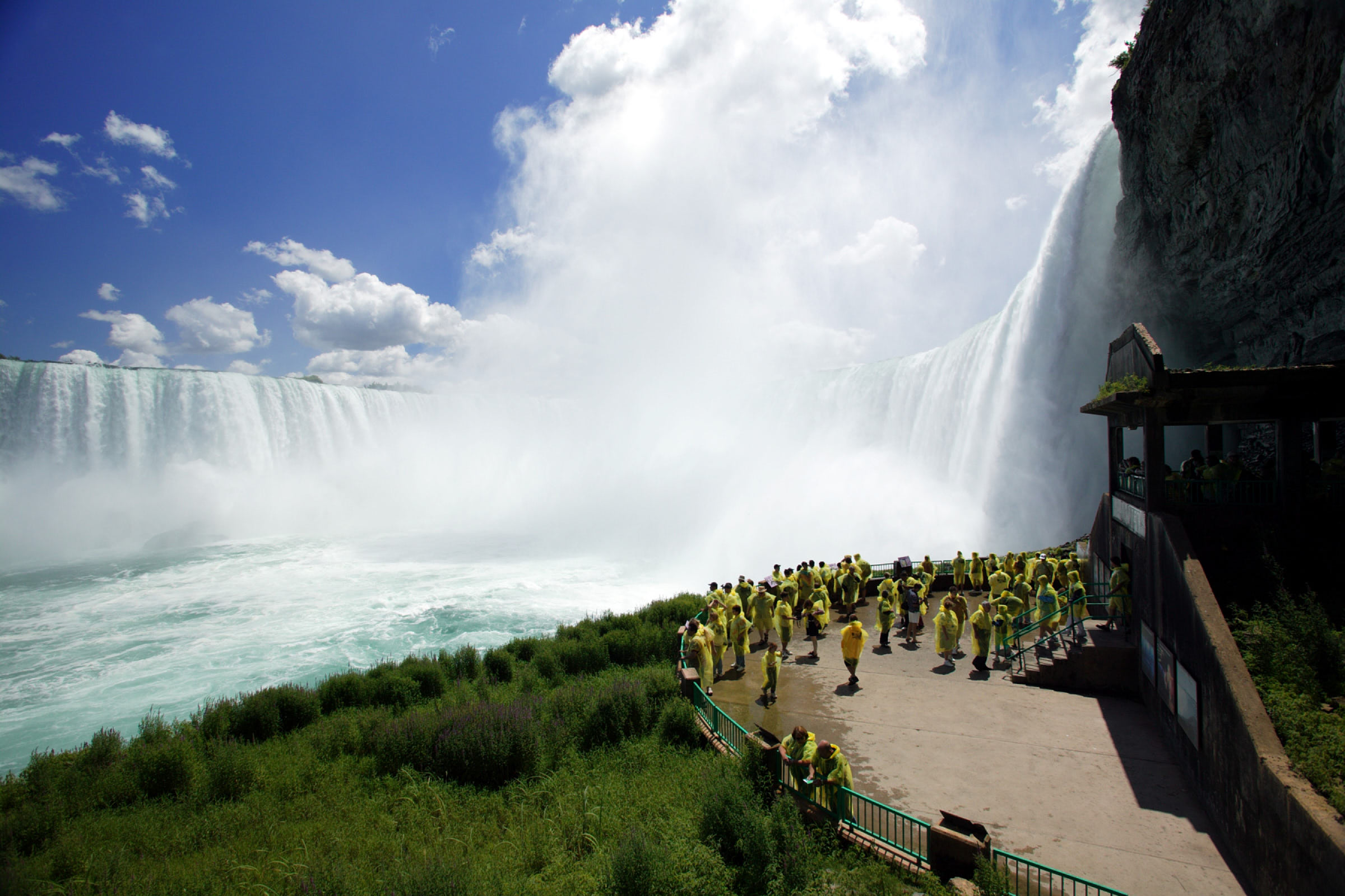 A midsize crowd wearing waterproof ponchos admire Niagara’s Falls Horseshoe Falls from lower observation deck on a sunny day