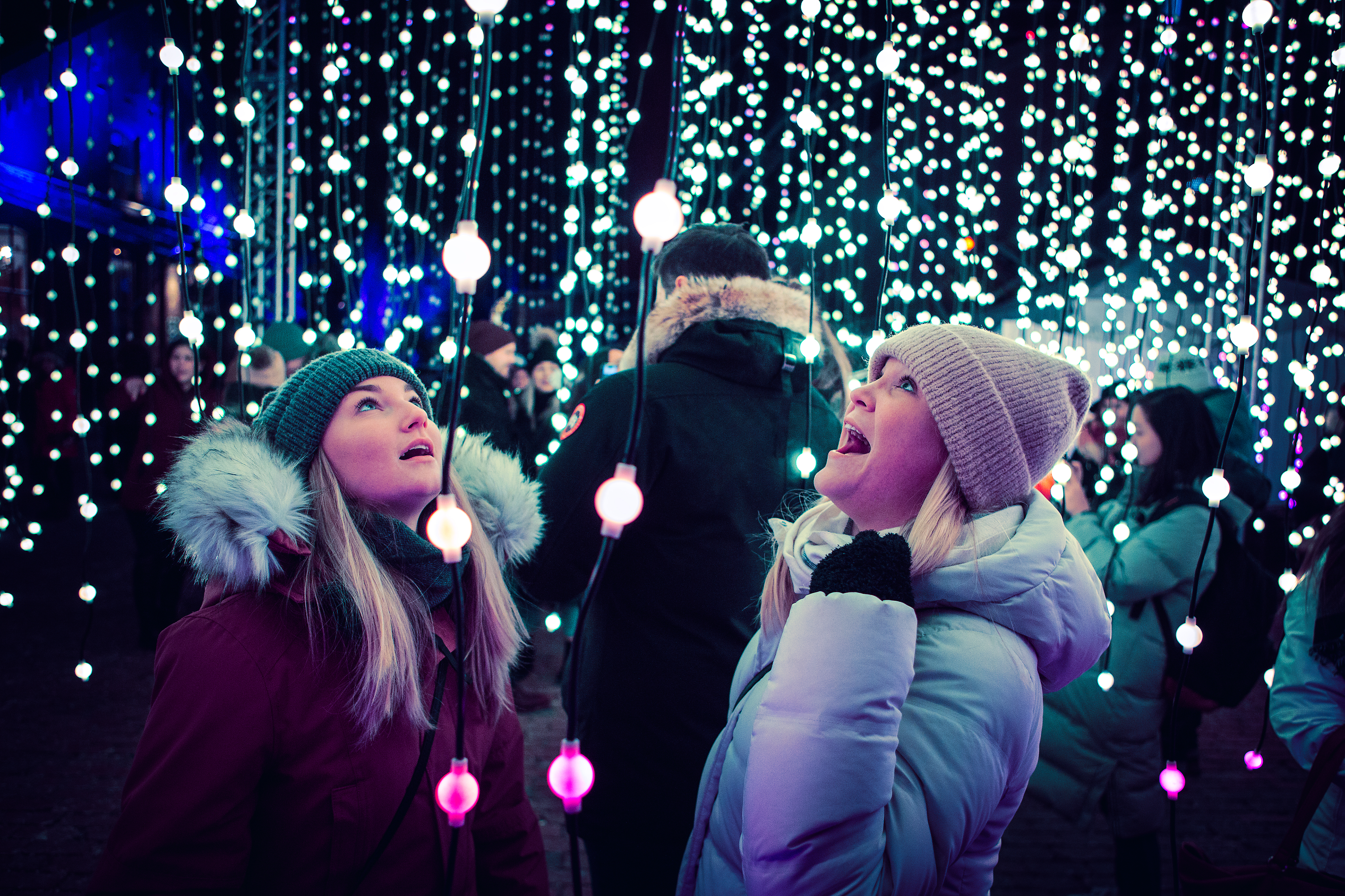 Two women in winter coats looking up at strings of lights at the Toronto Light Festival