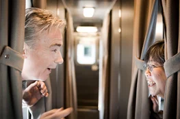 A man and women smile at eachother from the doorways of their cabins, across the train car corridor