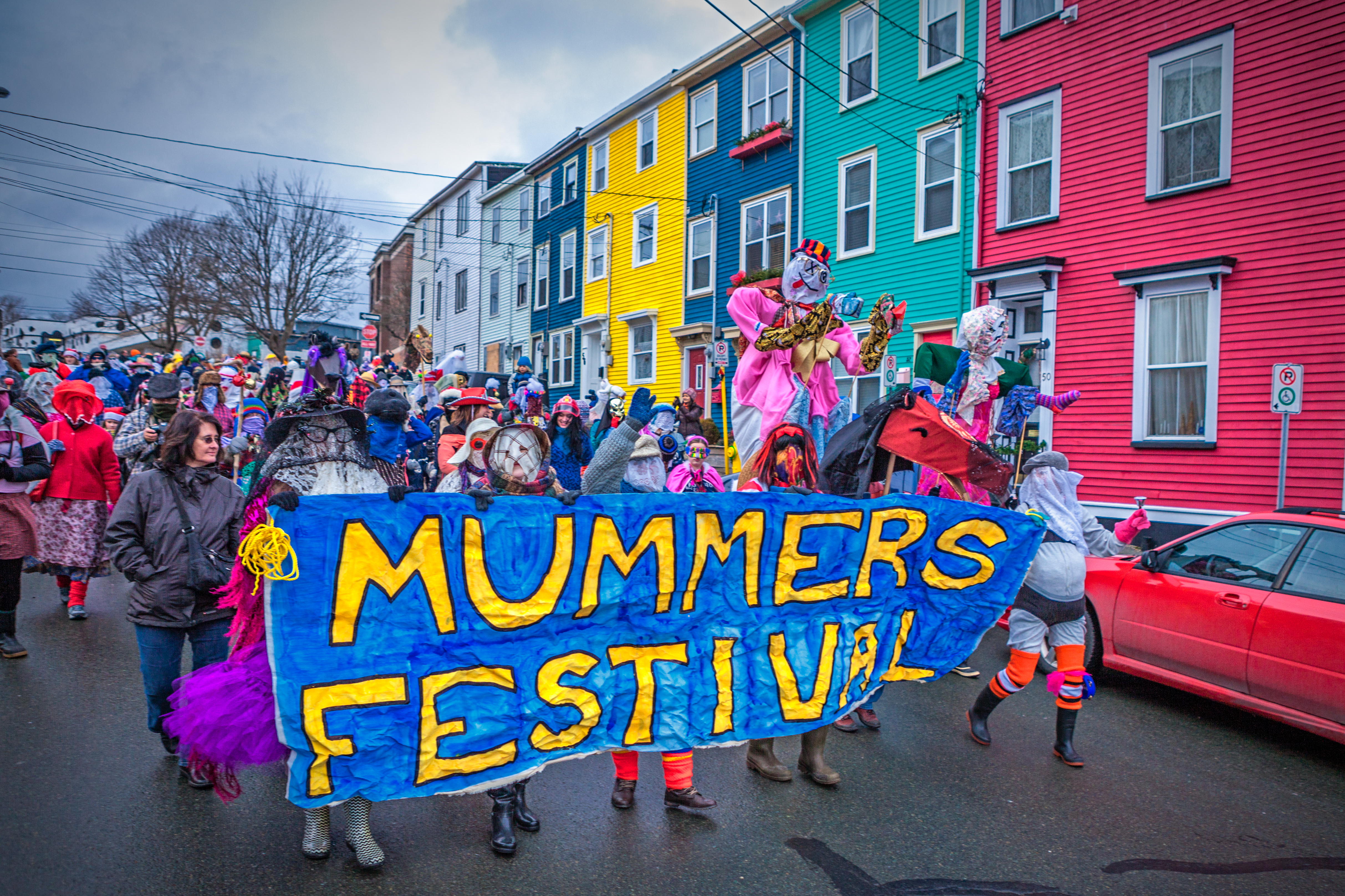 People holding a Mummers Festival sign in a parade past colourful houses in St. Johns