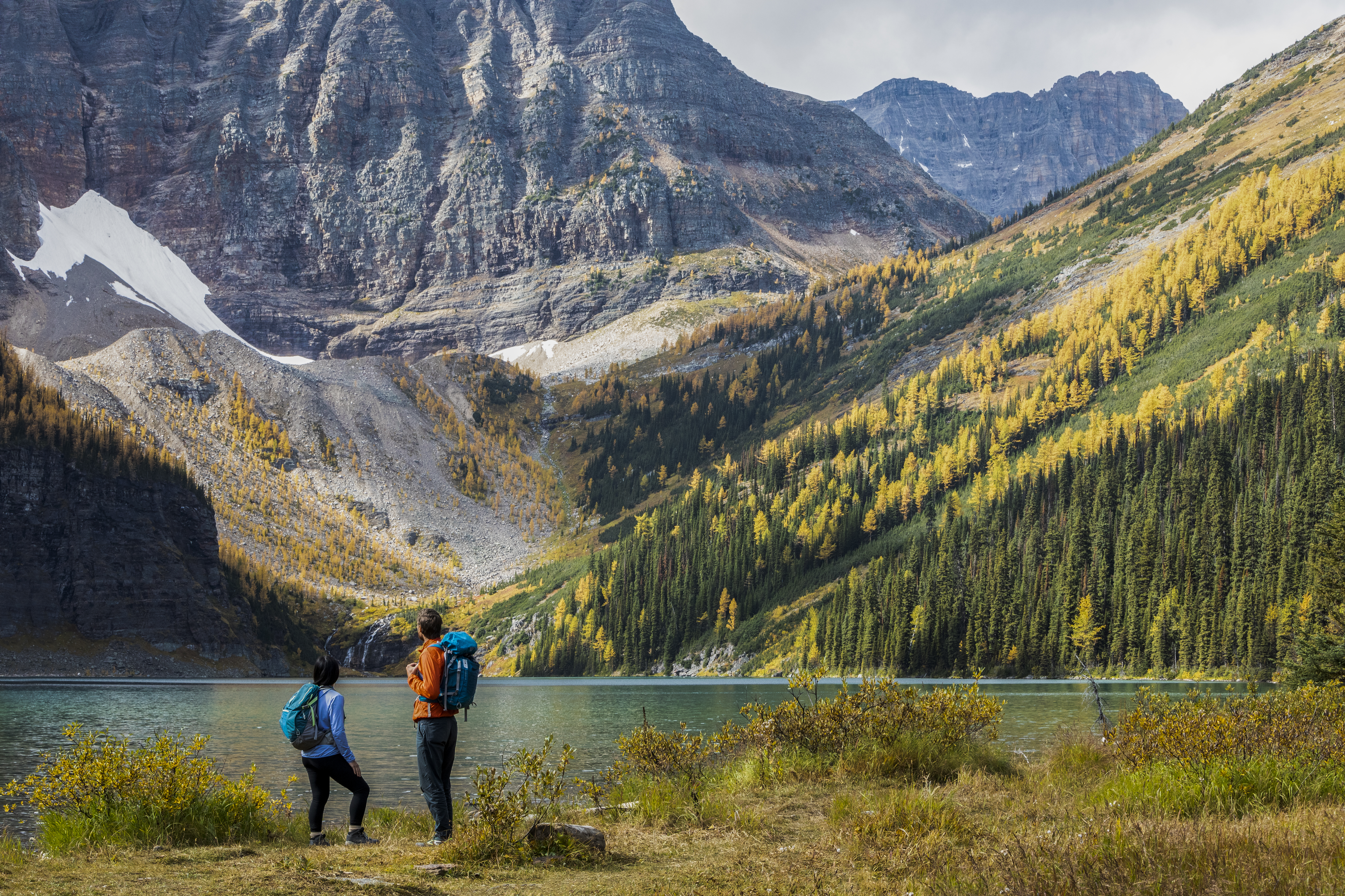Two visitors look ahead towards clear glacial lake, rolling mountains and thick forest leading to subalpine meadows located in Alberta's Lake Louise