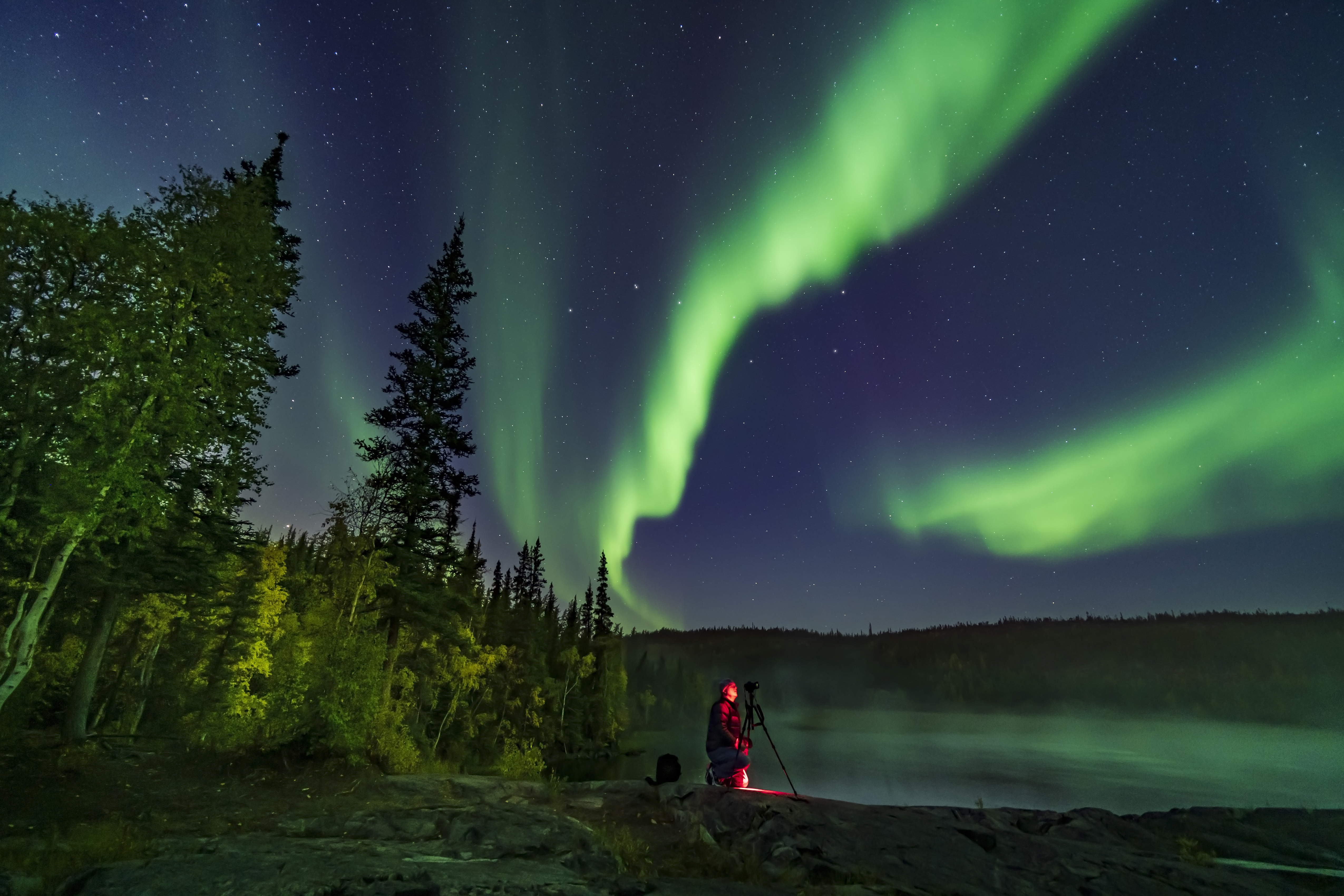 Man kneels behind camera on stand set up in Yellowknife on a day with vivid sighting of northern lights