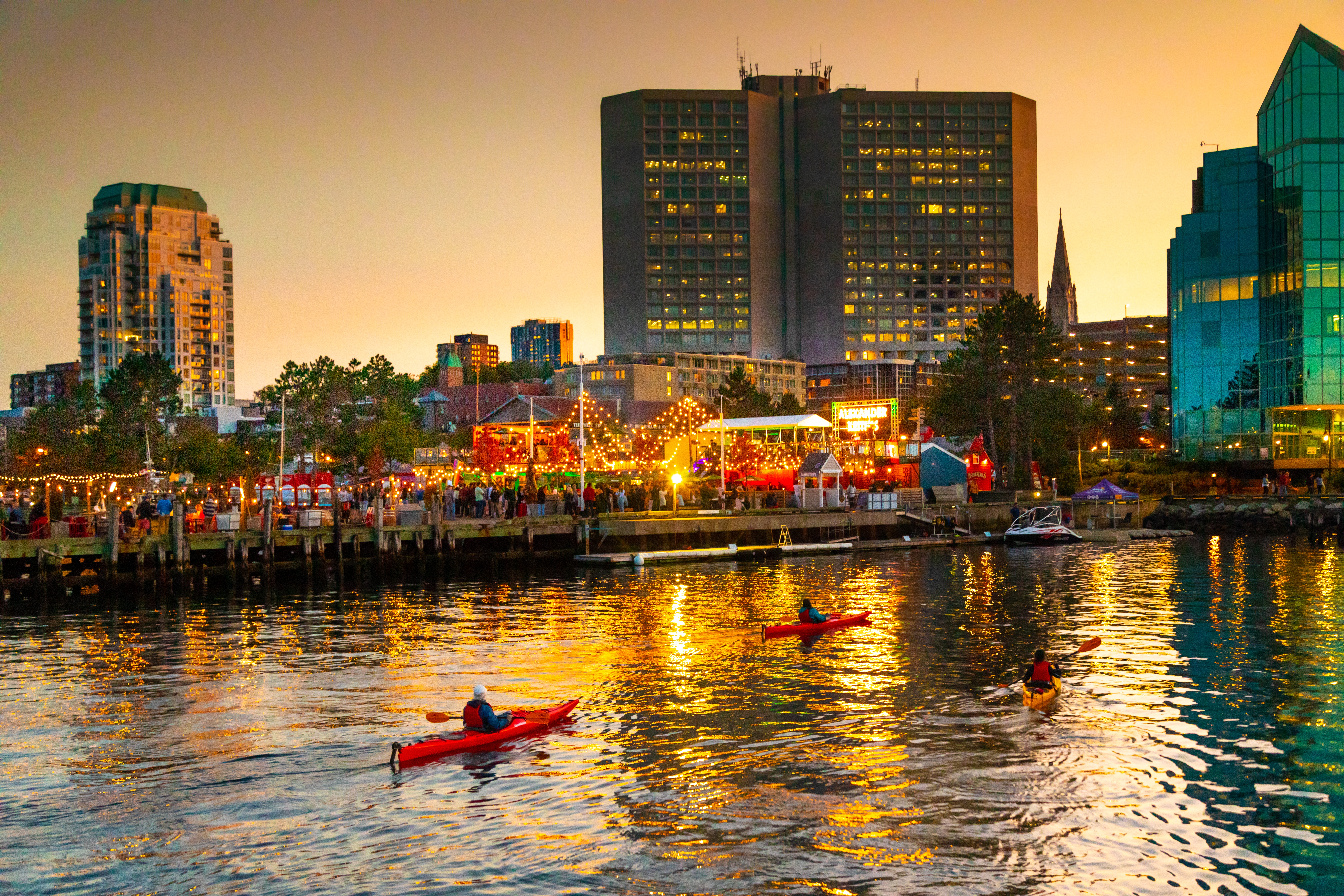 Sun sets as three kayakers pass by event with large attendance outdoors occurring by restaurant in Halifax Waterfront
