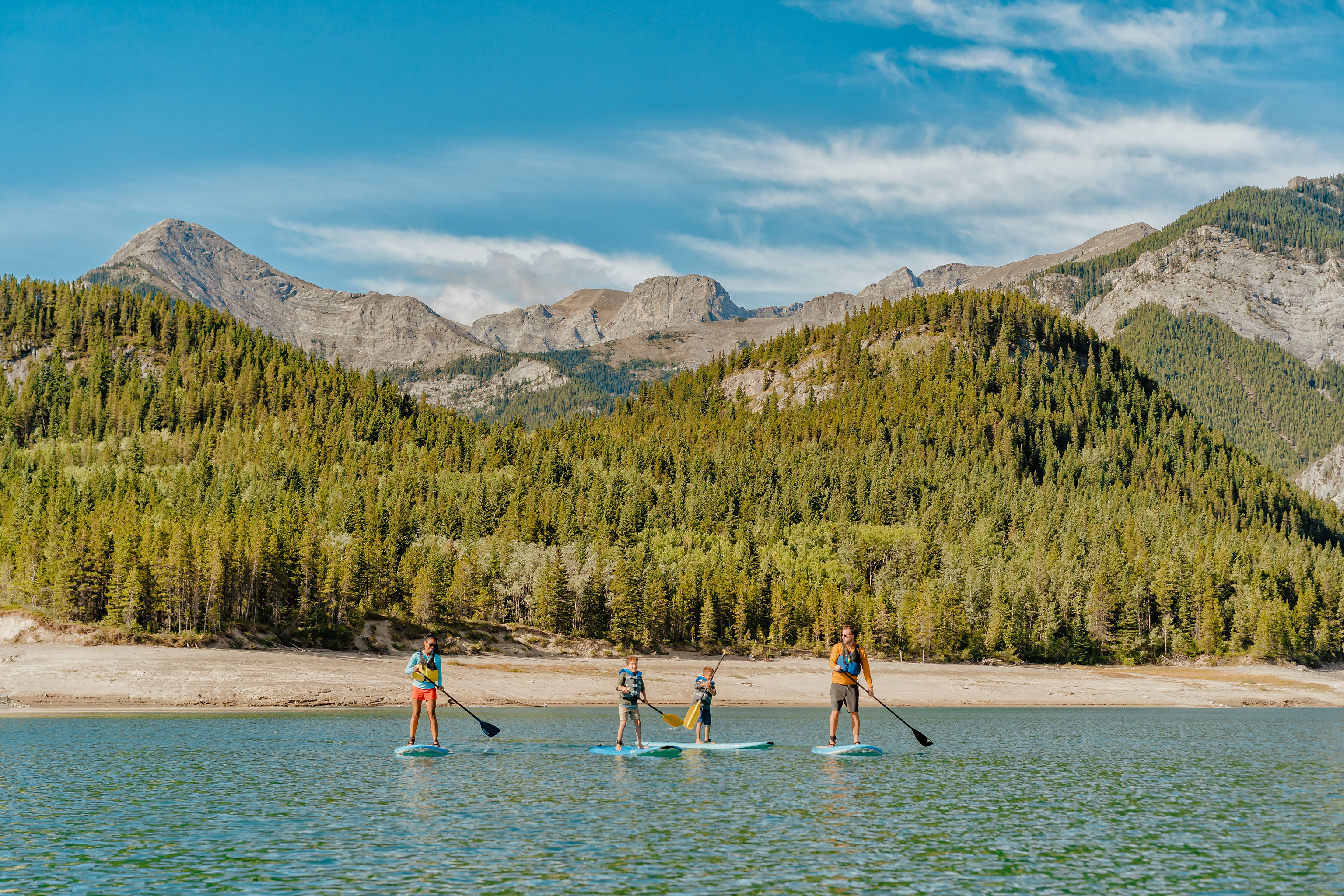 Four people stand-up paddleboard, by mountains, in Barrier Lake reservoir in Kananaskis' Bow Valley Provincial Park
