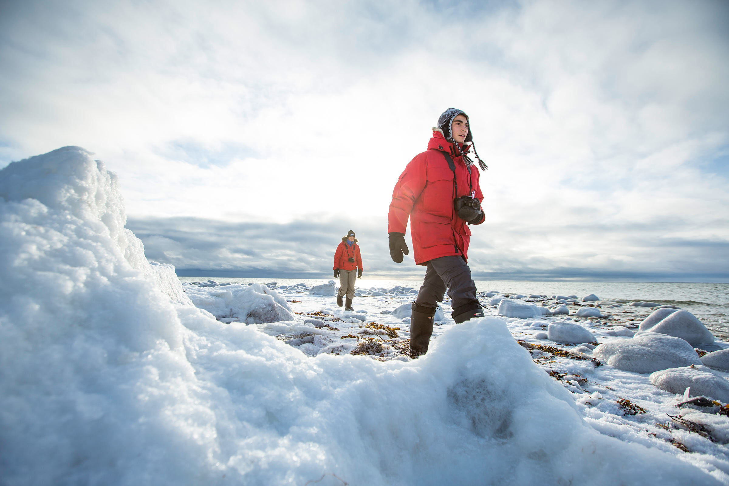 Two people in red winter jackets walk over ice and snow at Hudson Bay