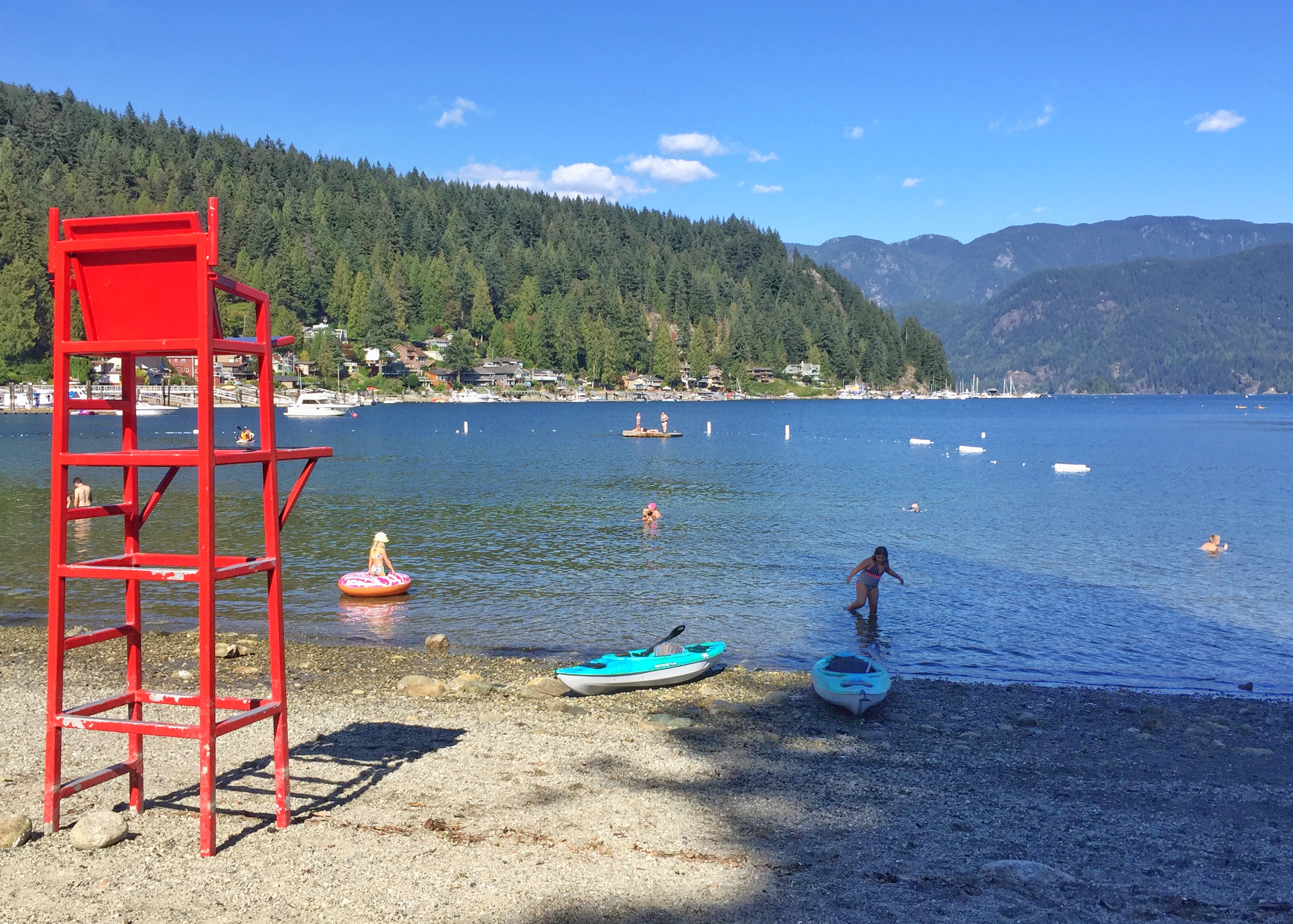 Kayaks and swimmers next to a red lifeguard chair on Deep Cove beach in summer