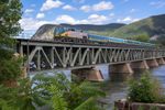 VIA Rail's Corridor train traveling on an iron bridge over water, with mountains in the background, framed by greenery.