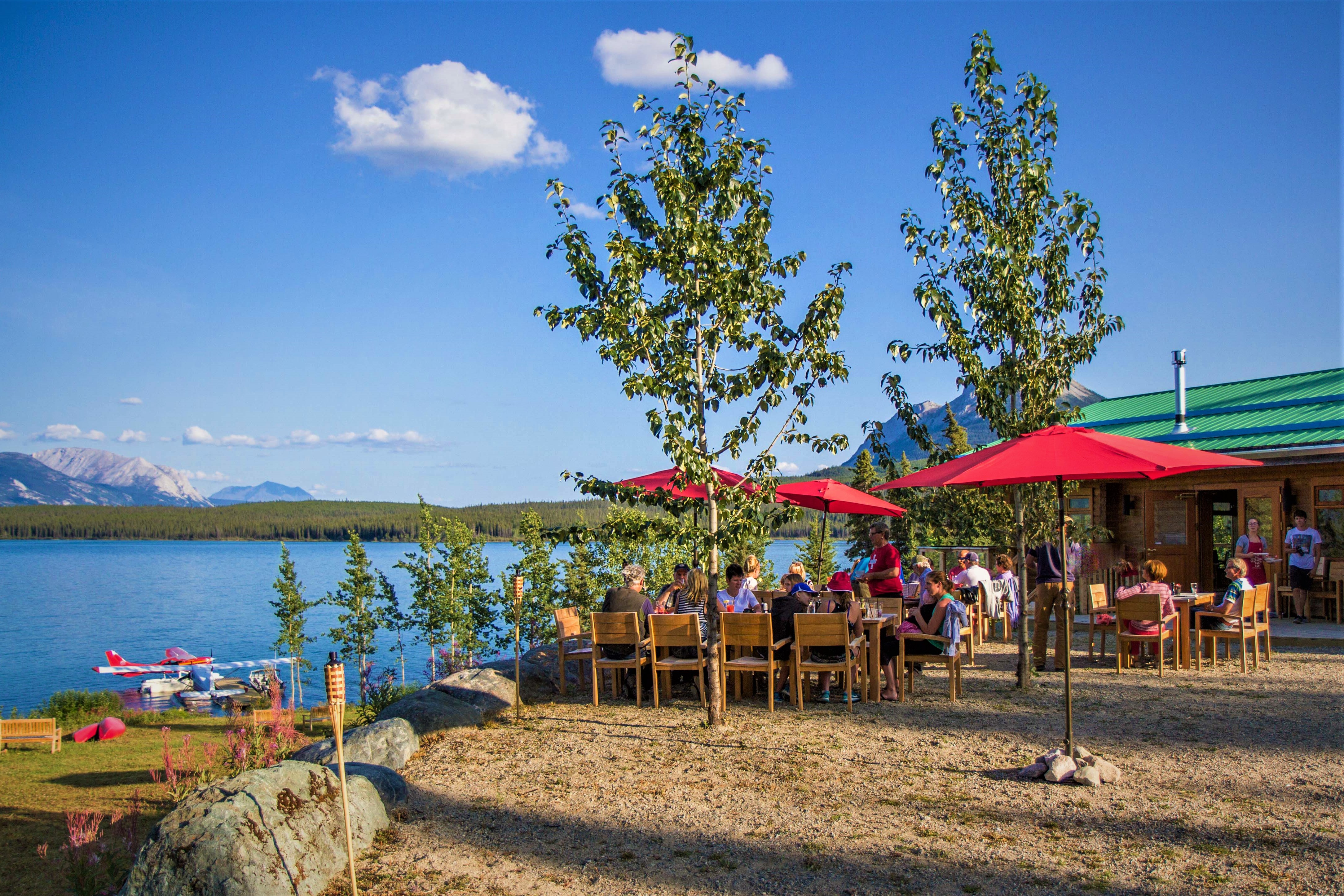 A group of people are sitting at tables under red umbrellas near a lake