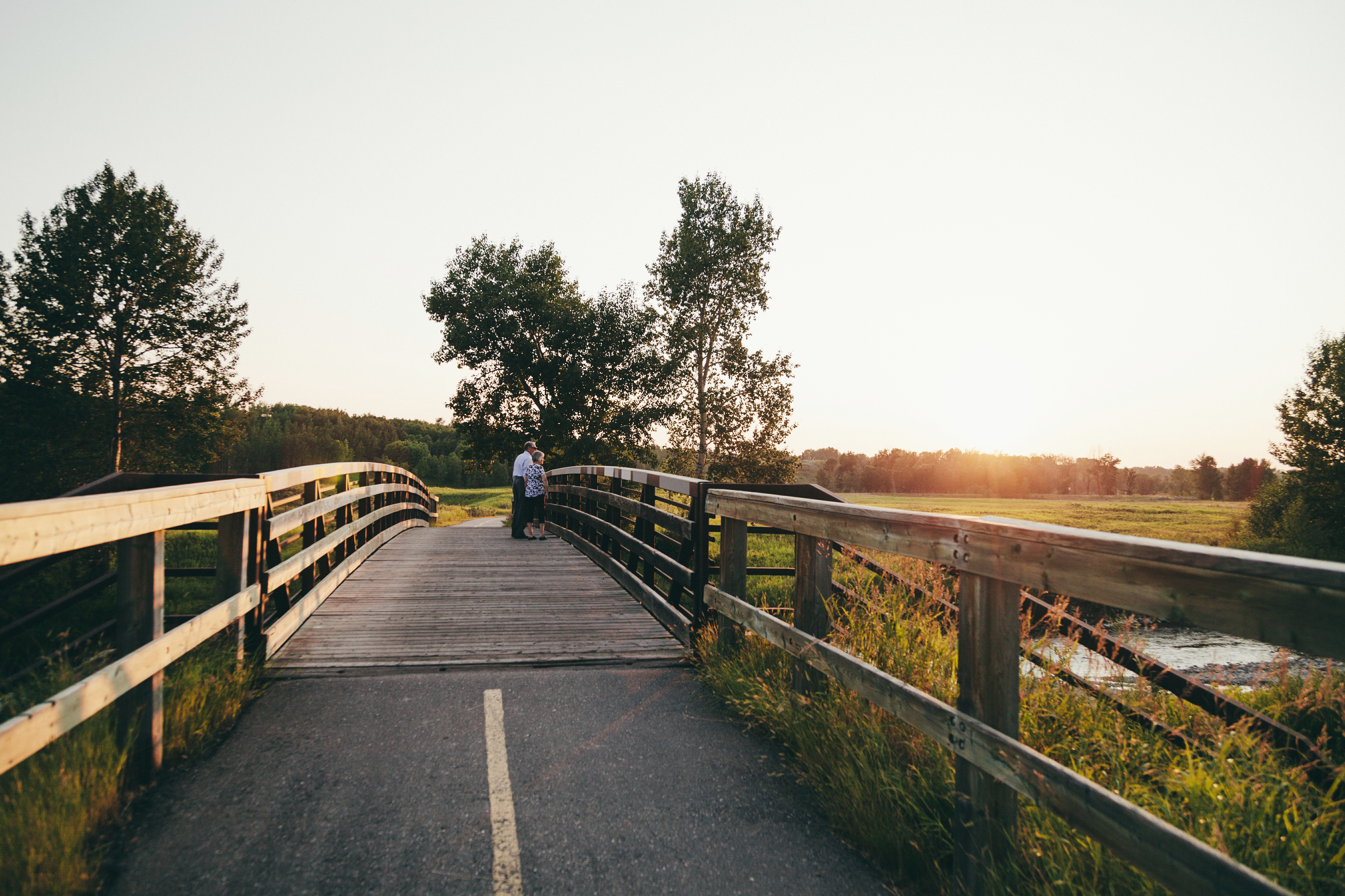 Couple stands on a bridge as the sun sets behind trees in Fish Creek Provincial Park