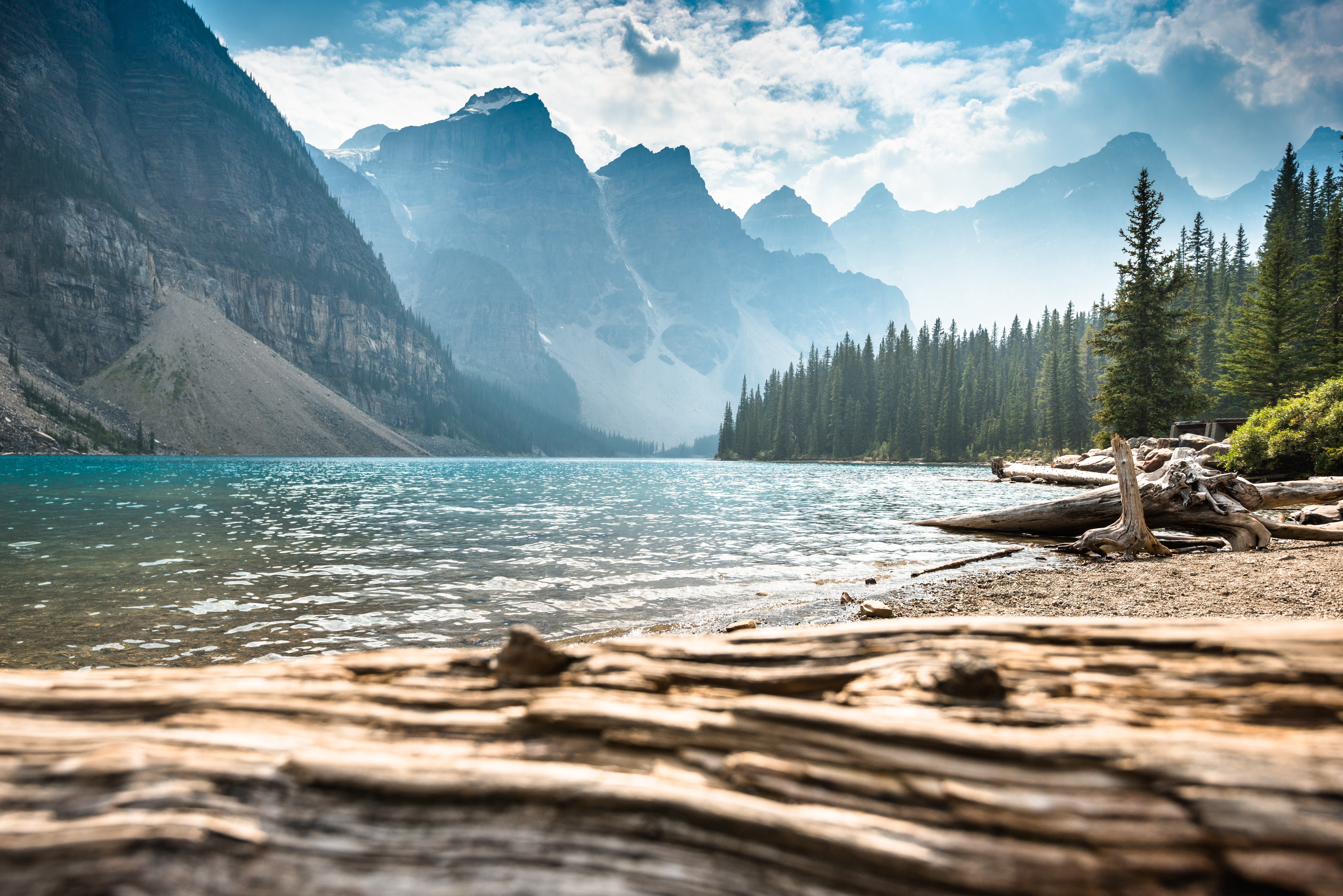 Moraine Lake clear waters glisten, under a bright sky, with row of Valley of the Ten Peaks mountains fading into a light blue shade and distance