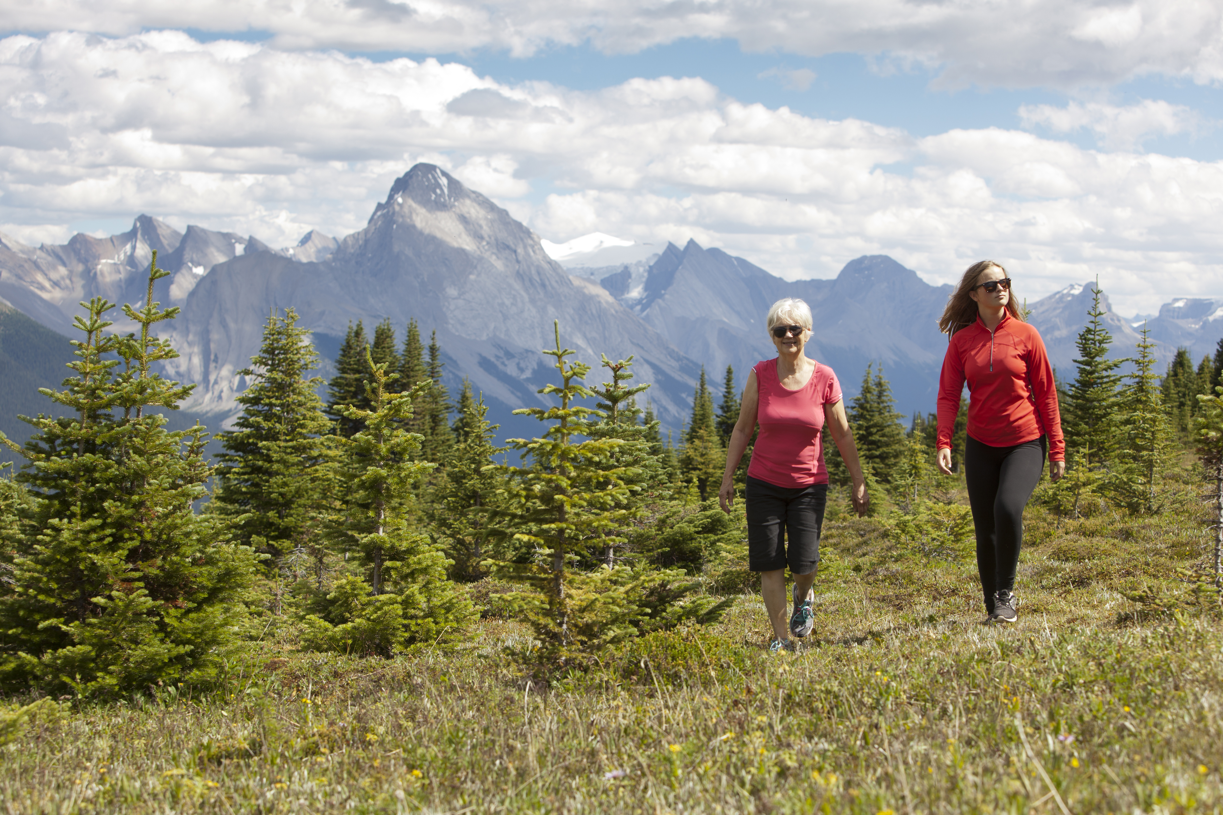 Two women walk in woods with jagged mountains in the distance