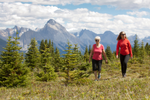 Two women hike through the forest in Jasper National Park with mountains in the background 