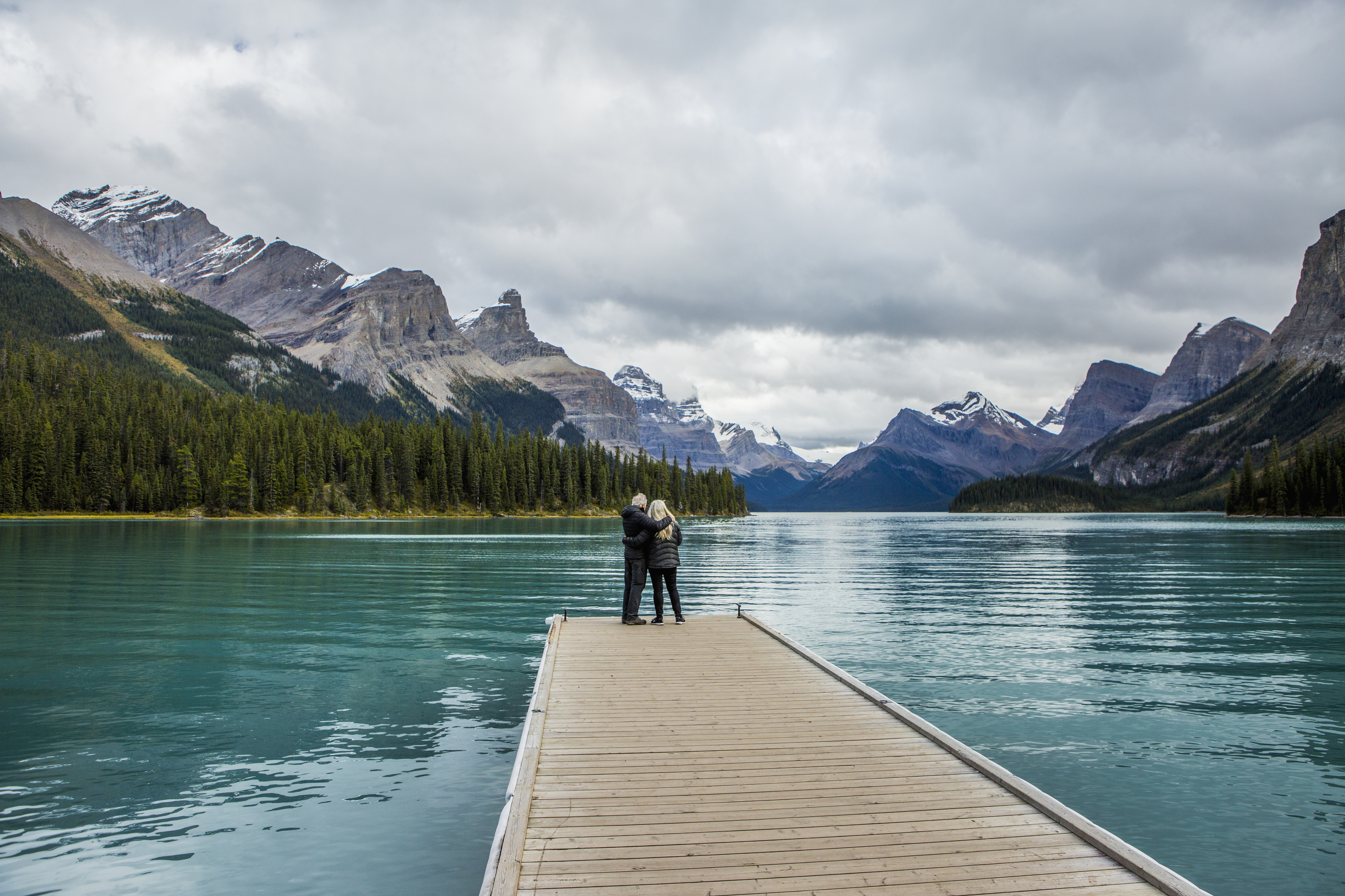 A senior couple stand at the end of a dock on Maligne Lake looking out at the turquoise water