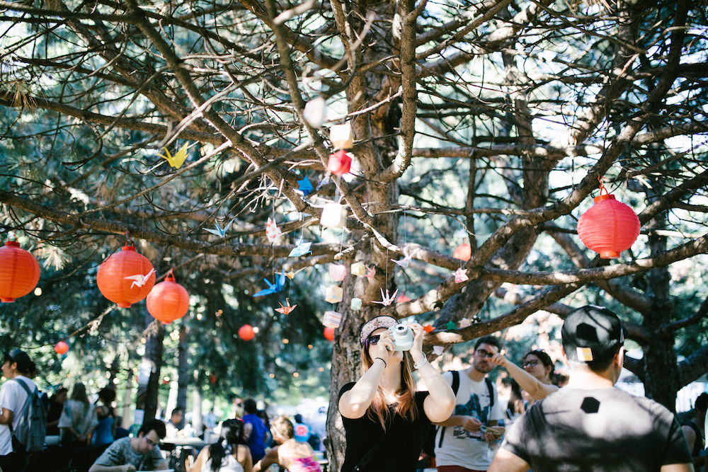 A girl takes a photo of a tree with origami cranes and paper lanterns hanging from it