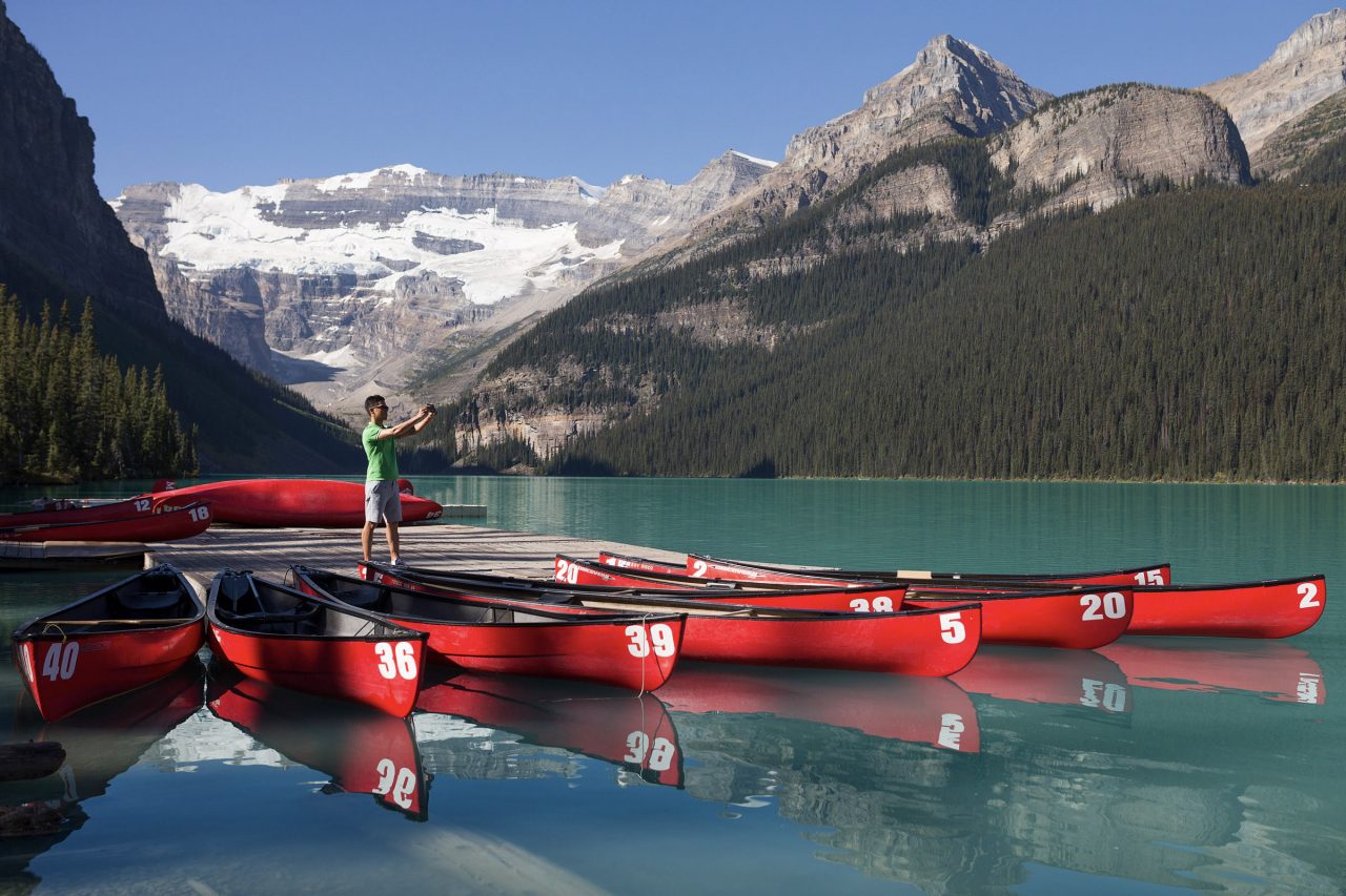 Man standing on a dock with red canoes with Lake Louise glacier behind