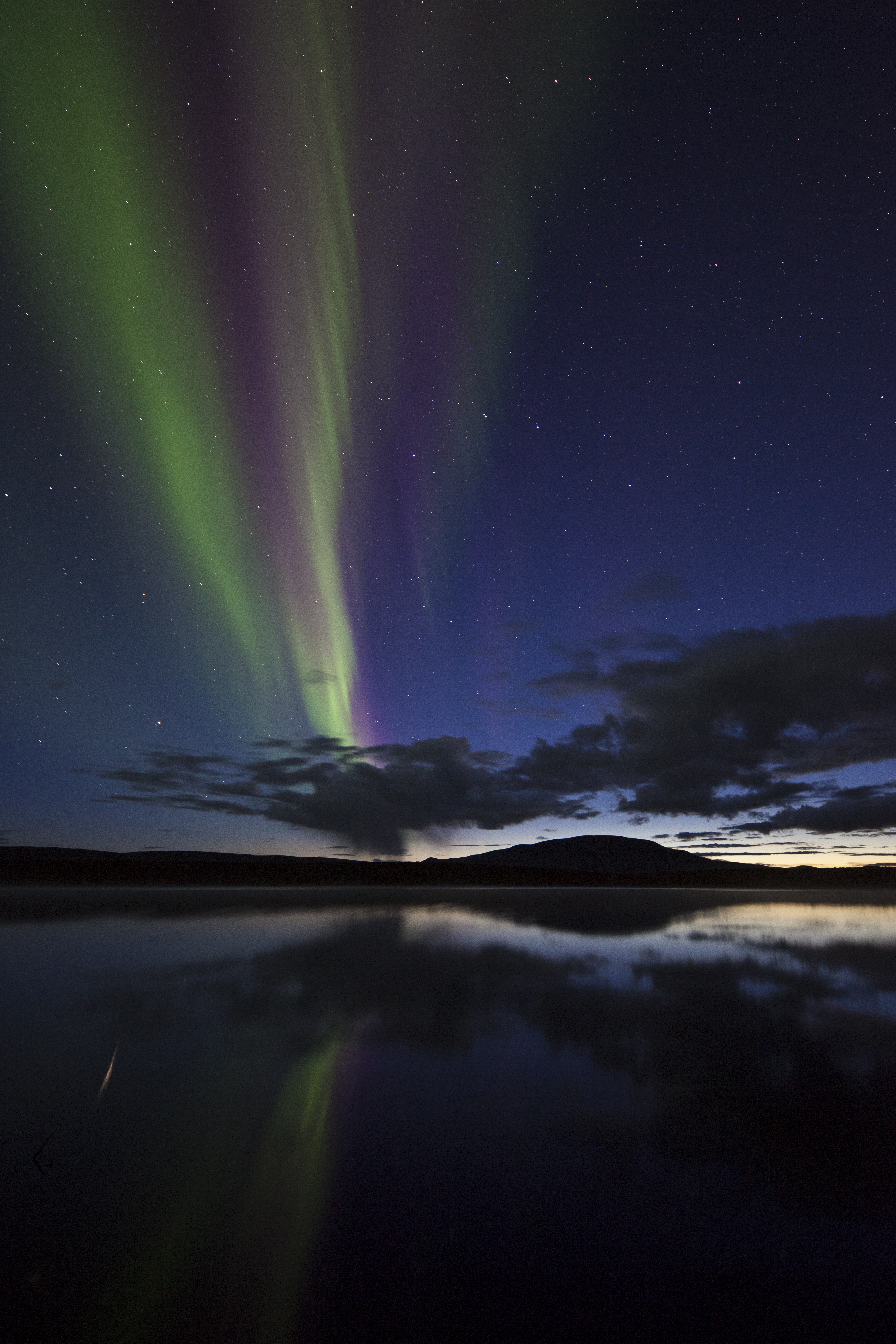 Aurora borealis appear across the sky like ribbons in the sky over horizon with vast body of water and silhouette of Mountains 