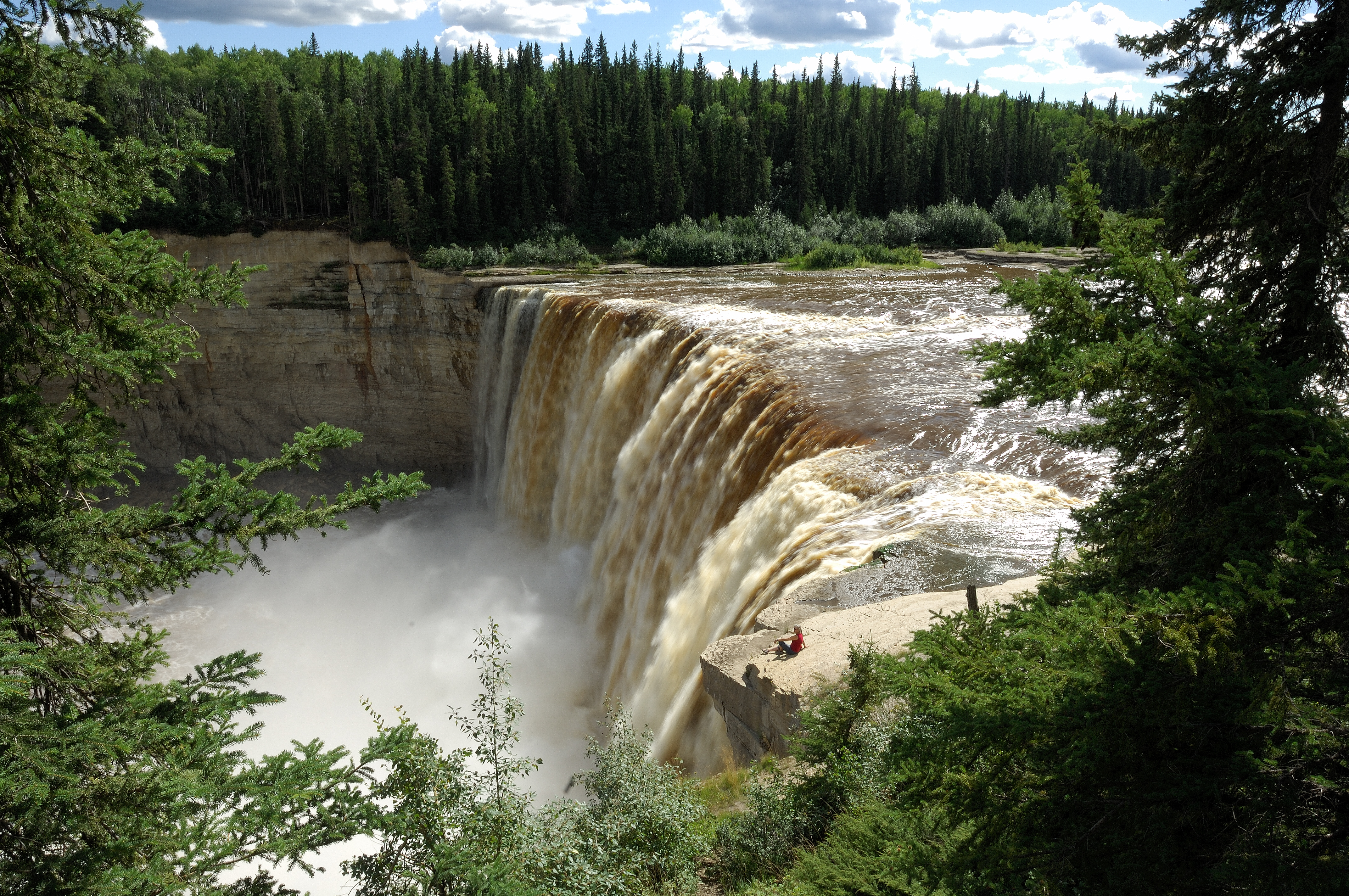 Person sitting on a rock next to a large rushing waterfall and river