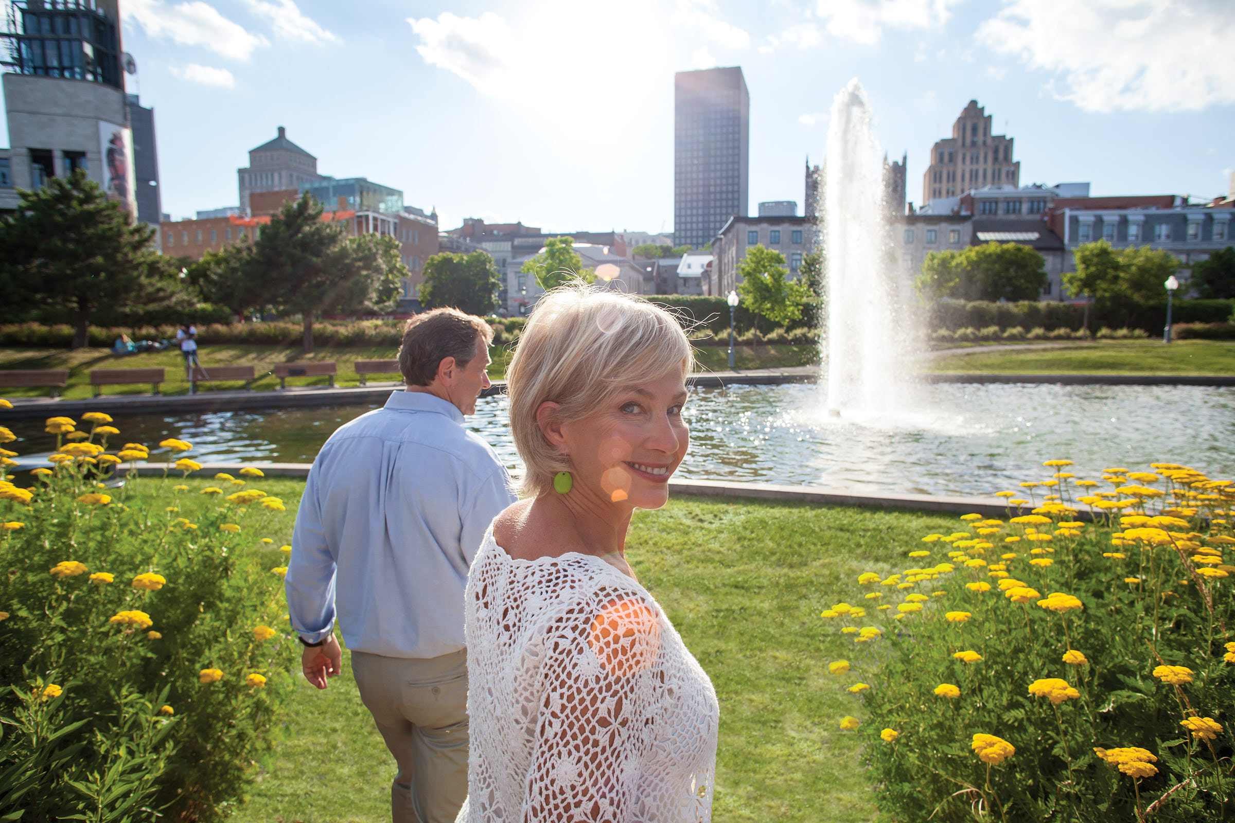 A senior woman looks at the camera as she walks with her partner towards a water feature in a garden
