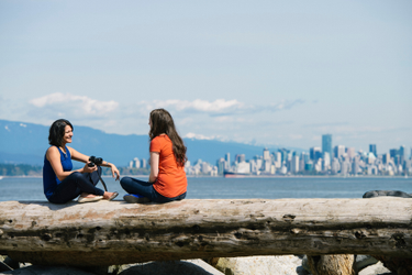 Two women sitting on a log at Jericho beach with the Vancouver skyline behind