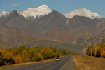 A car drives down the Alaska Highway with towering mountains in the distance and fall colours