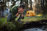 Man near yellow tent crouches on grass and splashes fresh water from stream of rushing water onto face in forested area in Banff National Park Campground